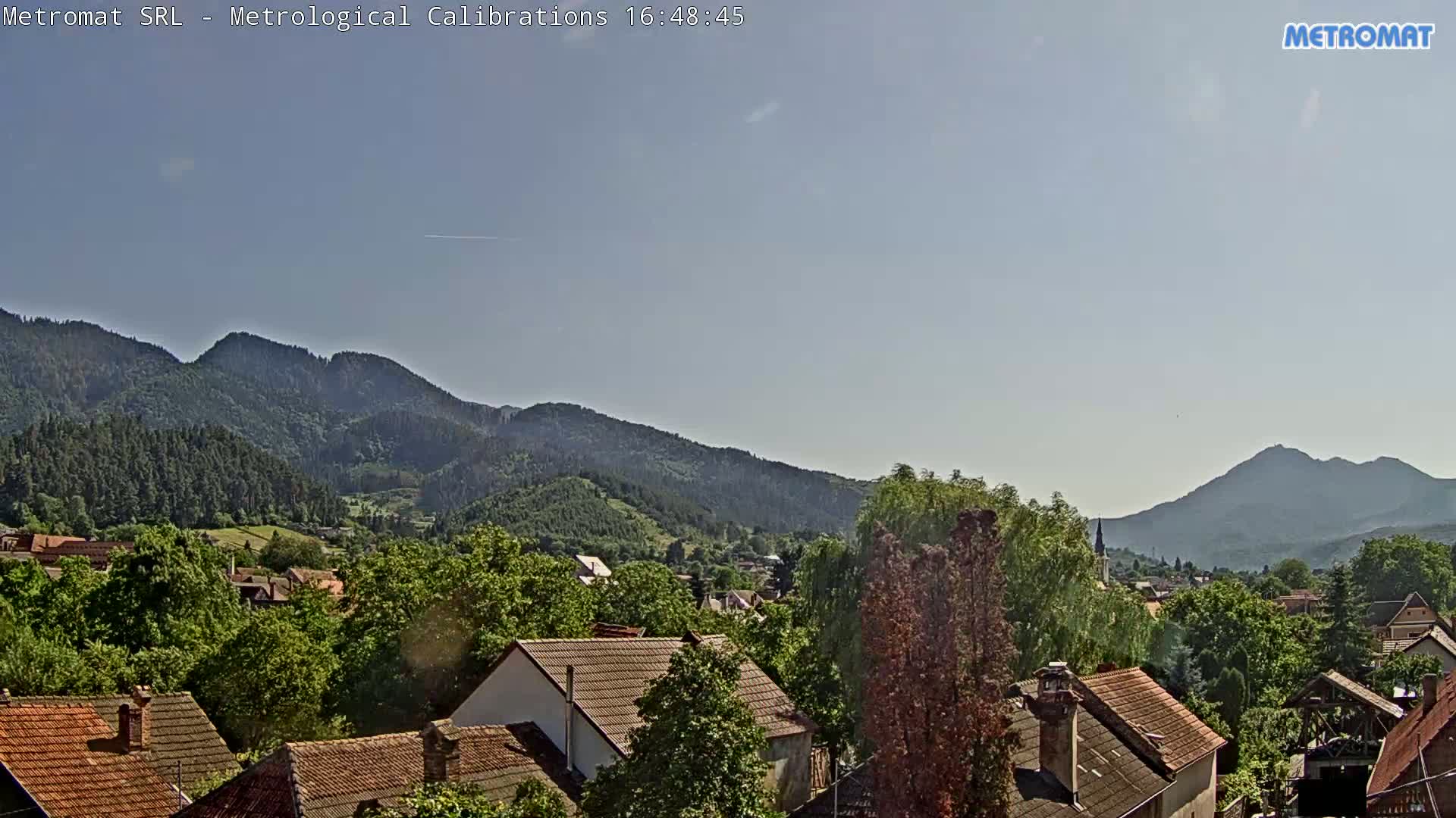 A clear, sunny day reveals a village nestled amongst lush green trees and mountains.