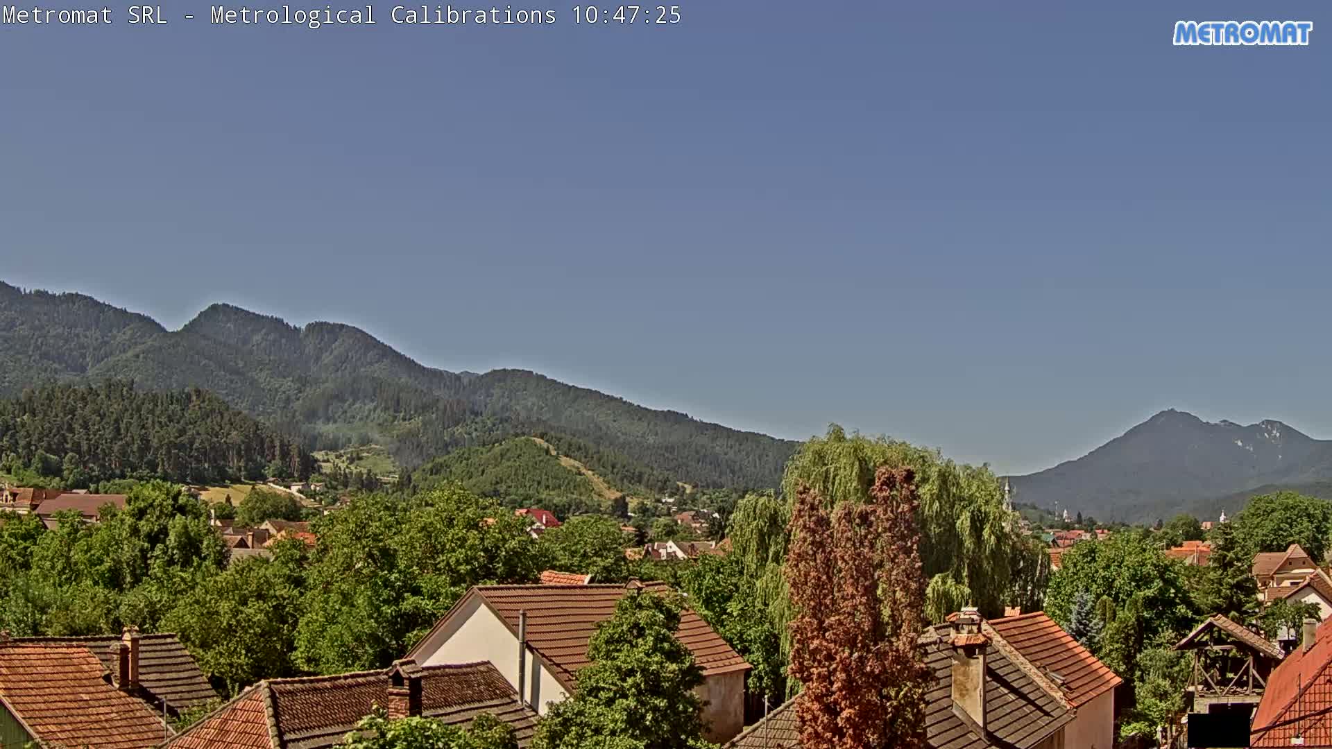 A small town with red-tiled roofs nestles amongst lush green trees and hills under a clear blue sky.
