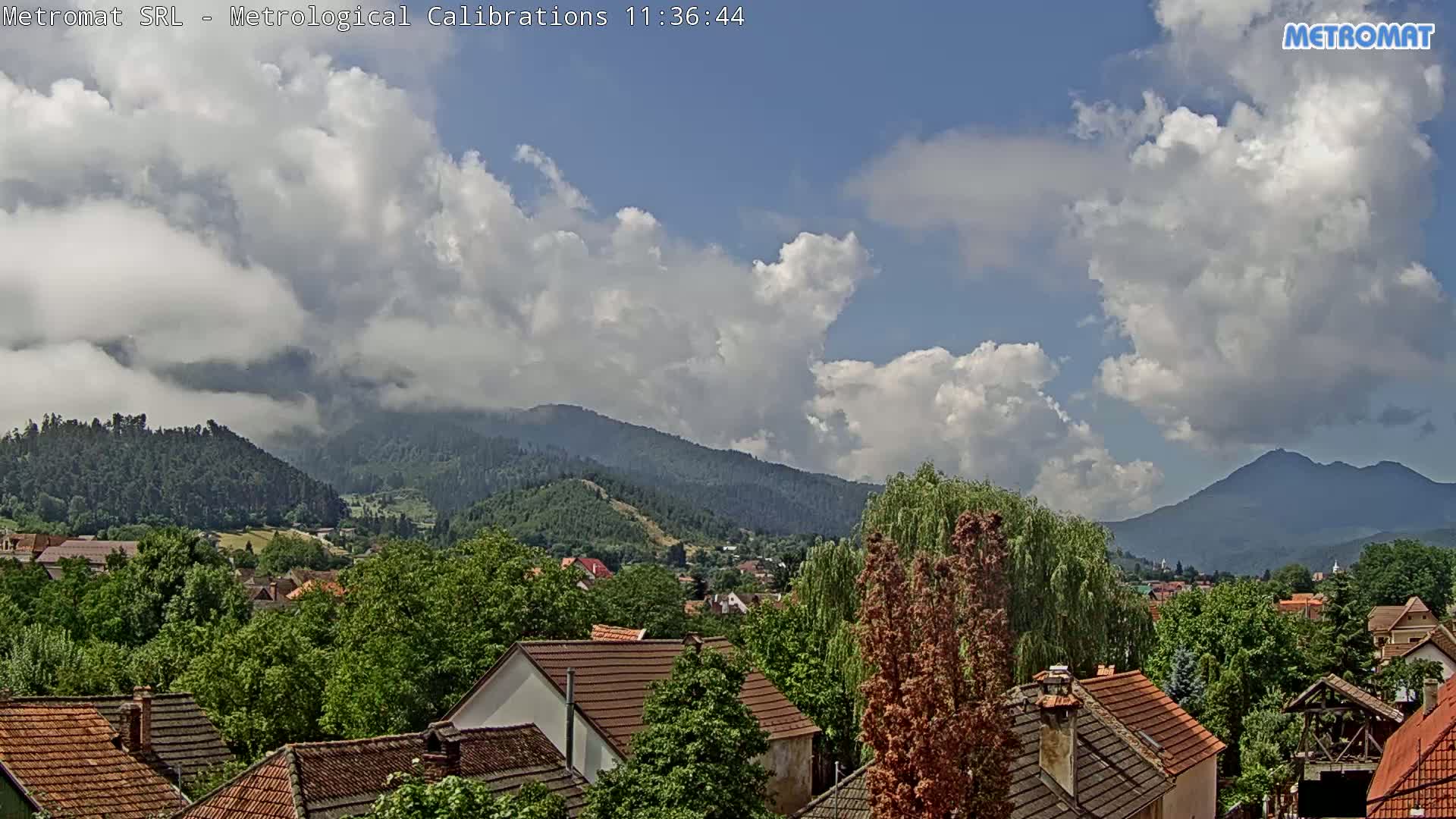 A partly cloudy sky overlooks a town nestled among green, tree-covered hills and mountains.
