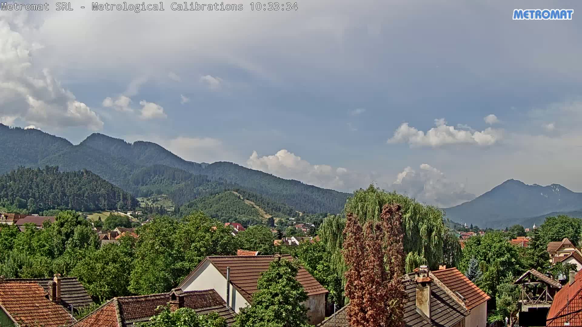 A partly cloudy sky overlooks a village nestled among green, tree-covered hills and mountains.