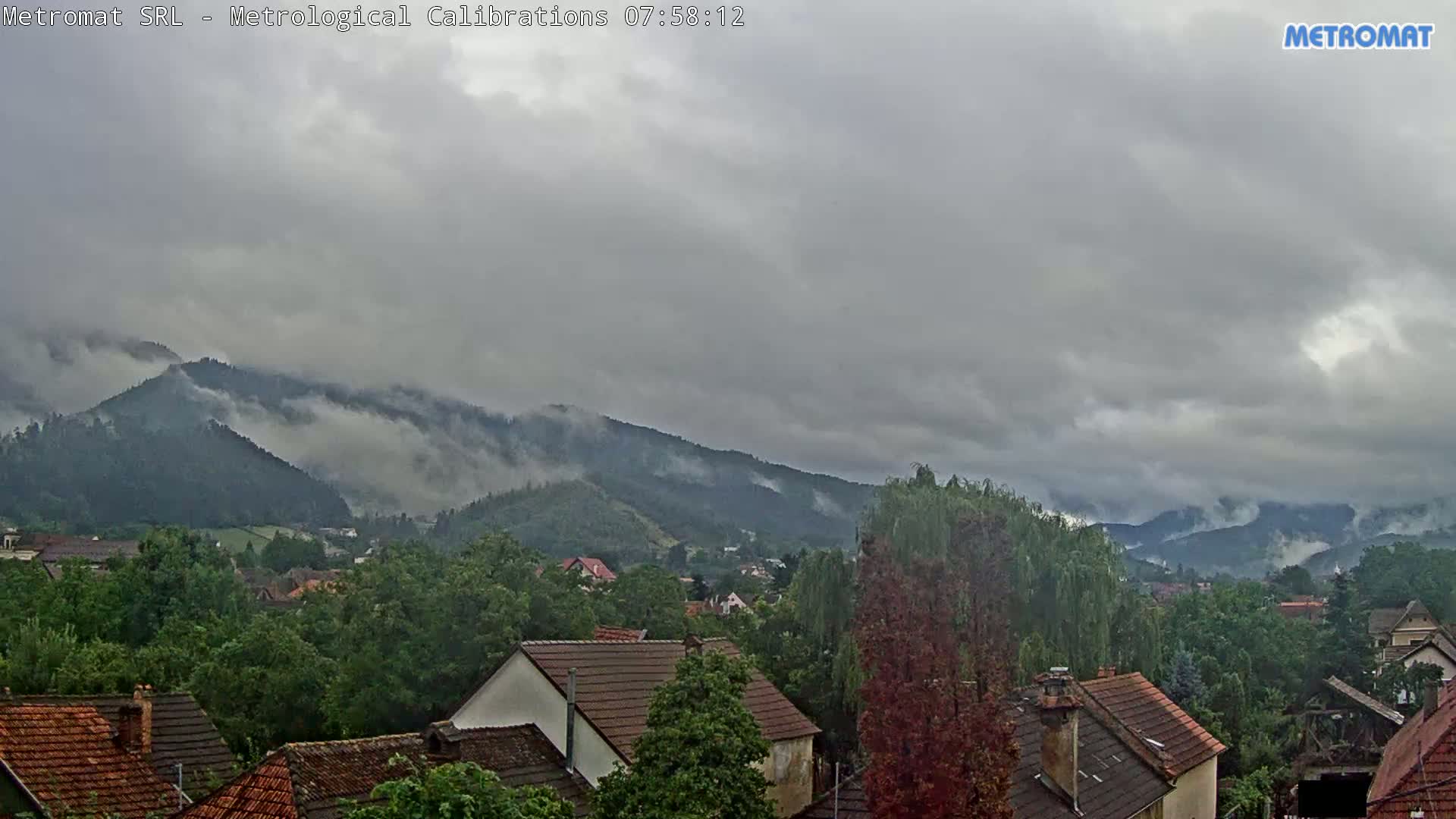 A partly cloudy sky overlooks a town nestled among green, tree-covered mountains.