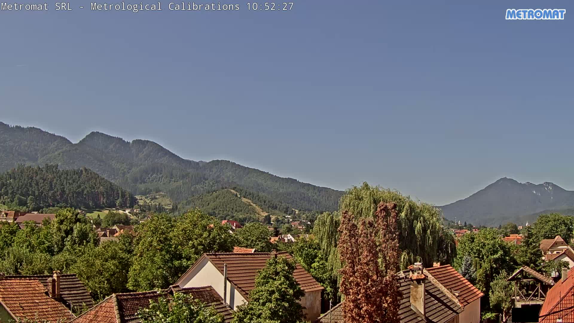 A clear, sunny day reveals a village nestled among green trees and mountains.