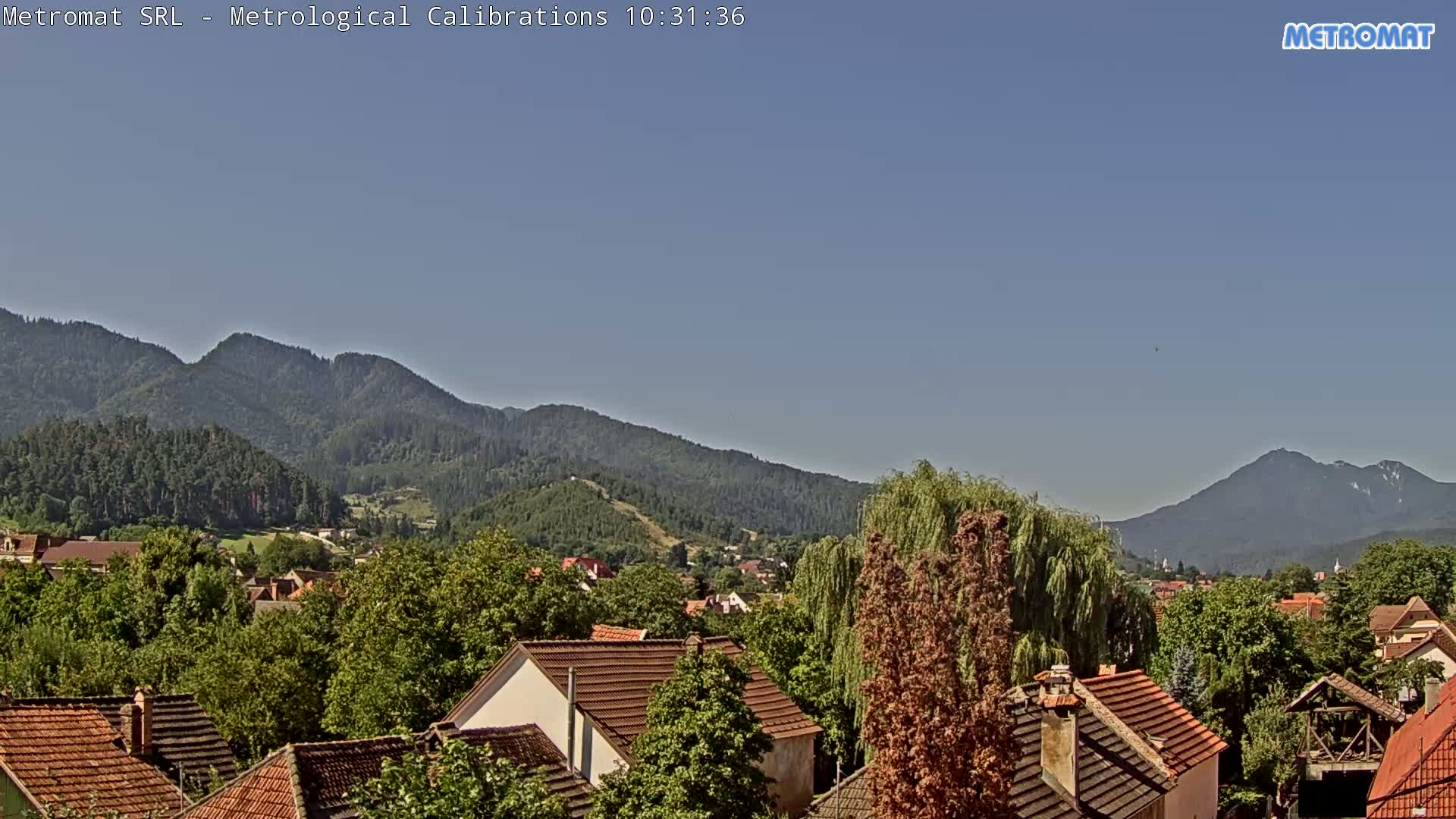A clear, sunny day reveals a small town nestled among green trees and mountains.