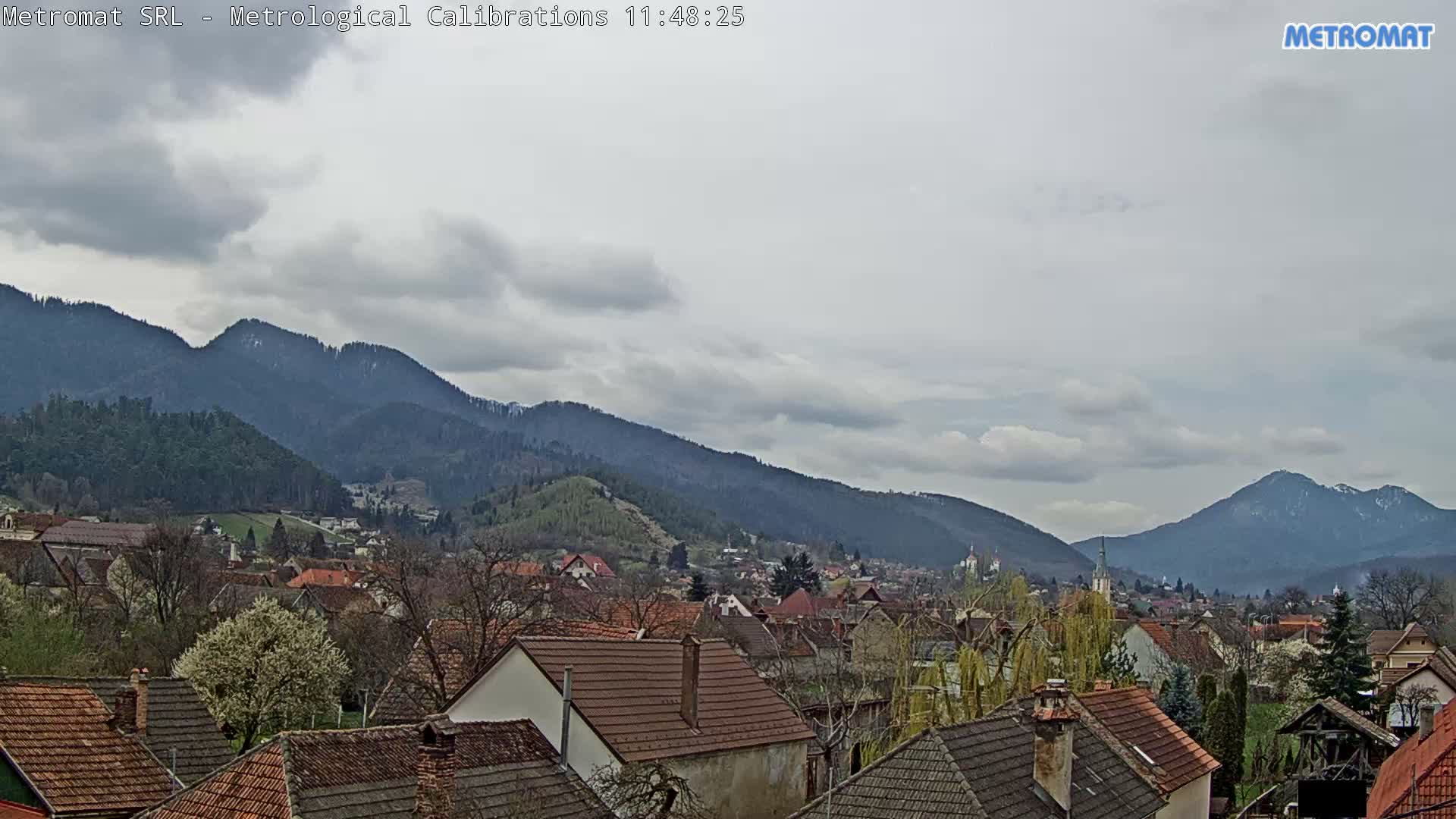 A village with numerous bare trees is nestled in a valley between forested mountains, all under a heavy, overcast sky.
