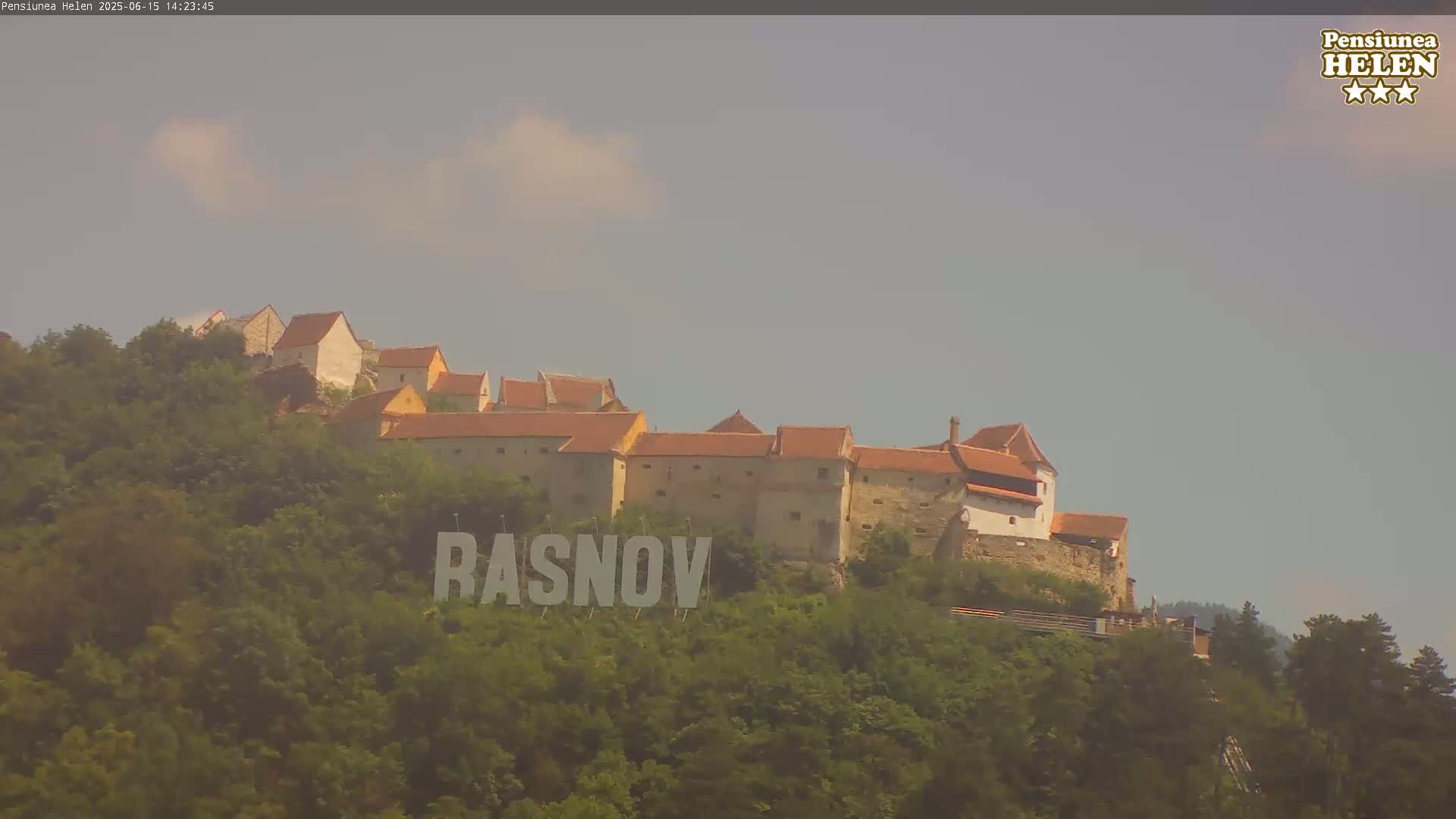 A hilltop castle with red-tiled roofs is nestled among green trees under a hazy sky.