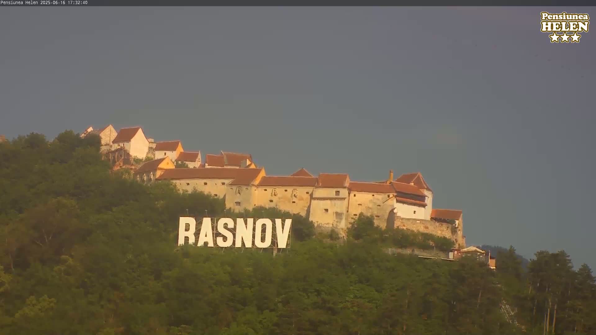 A hilltop castle with orange-tiled roofs is situated under a cloudy sky, and a large sign reading "RASNOV" is visible at the base of the hill.