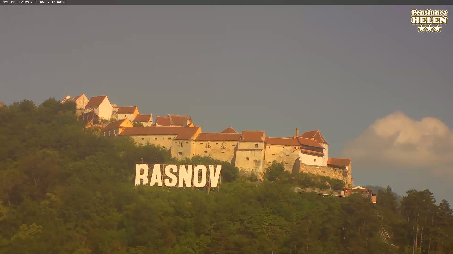 A large stone castle sits atop a hill covered in green trees, with a large sign reading "RASNOV" in the foreground under an overcast sky.