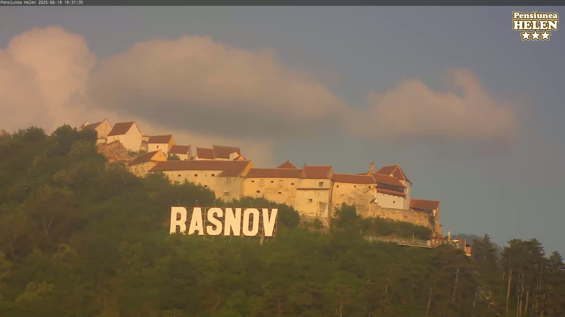 A hilltop castle with red-tiled roofs is seen under a partly cloudy sky, with a large sign reading "RASNOV" in the foreground.