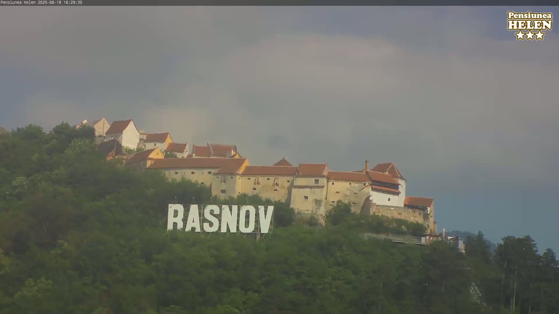A large sign reading "RASNOV" sits in front of a hilltop castle under an overcast sky.