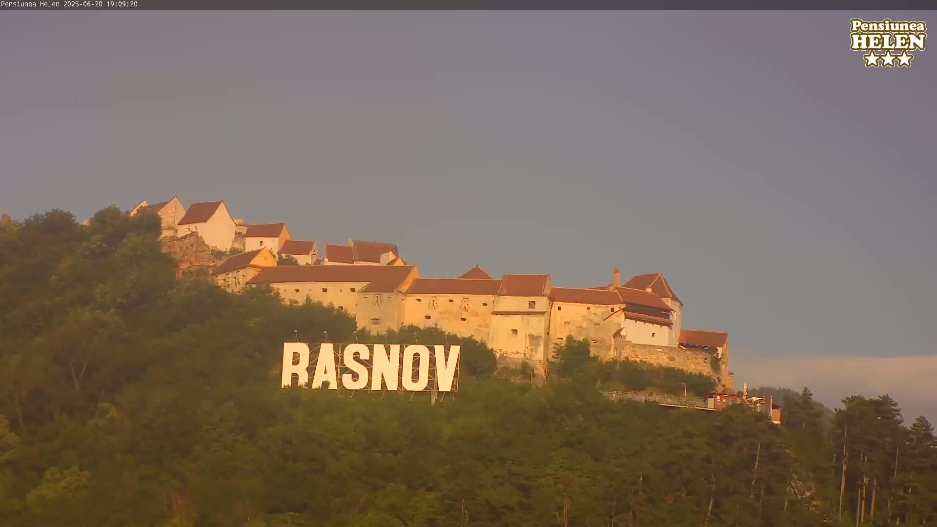 A large castle sits atop a hill covered in green trees, under a hazy, light-grey sky, with a large sign reading "RASNOV" in the foreground.