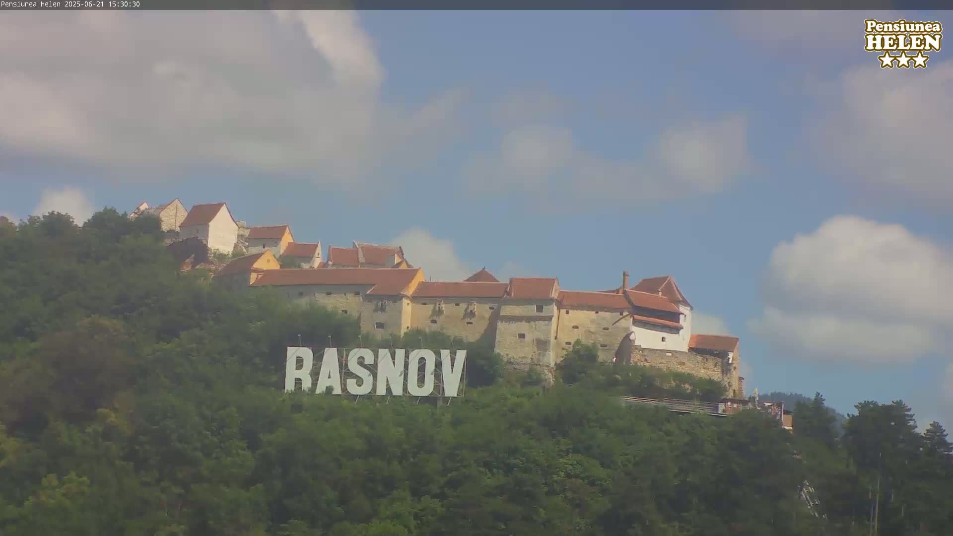 A large stone castle sits atop a hill covered in lush green trees under a partly cloudy sky.