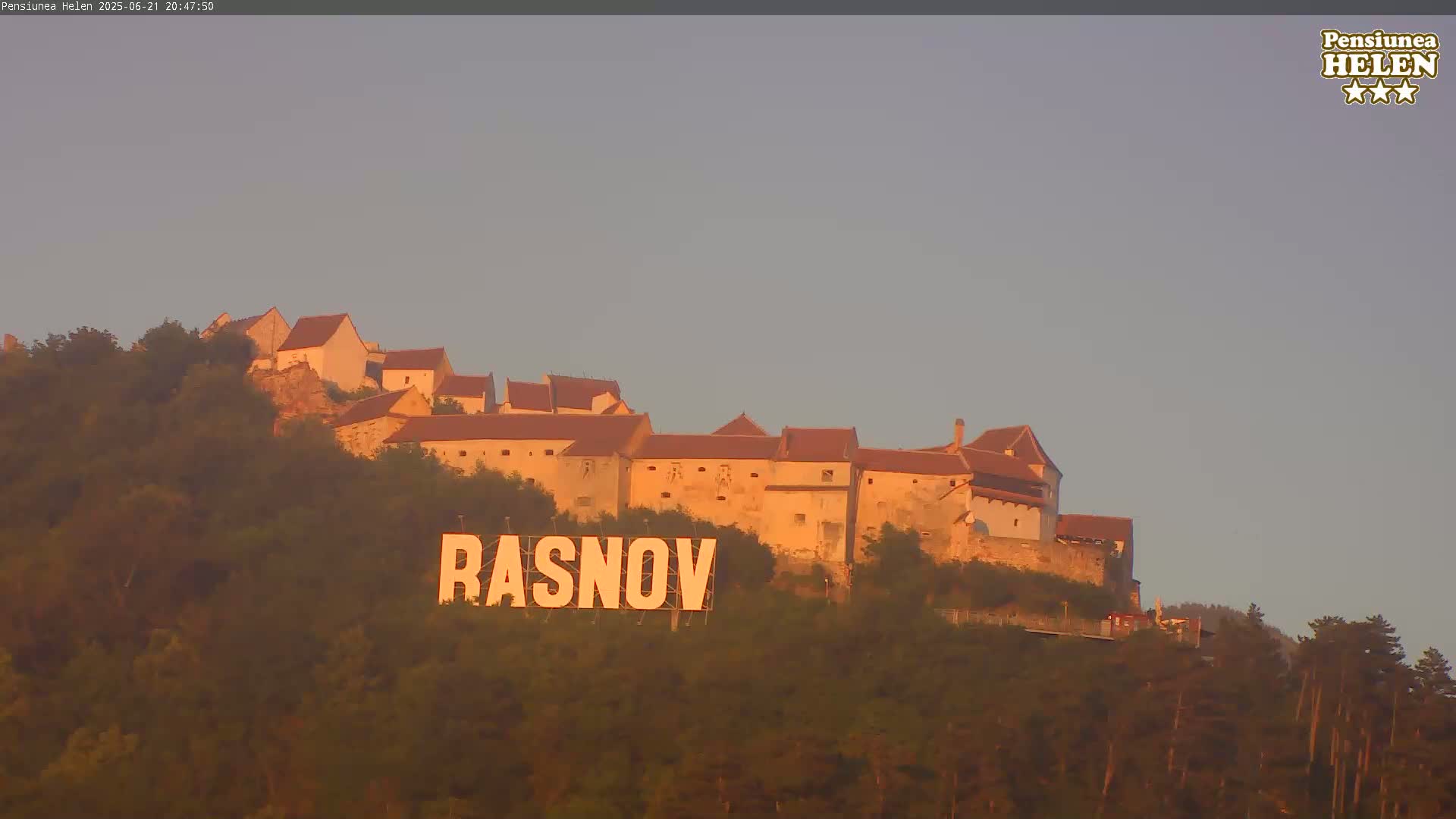 A hilltop castle is bathed in the warm light of a setting sun, with a large sign reading "RASNOV" prominently displayed below it.