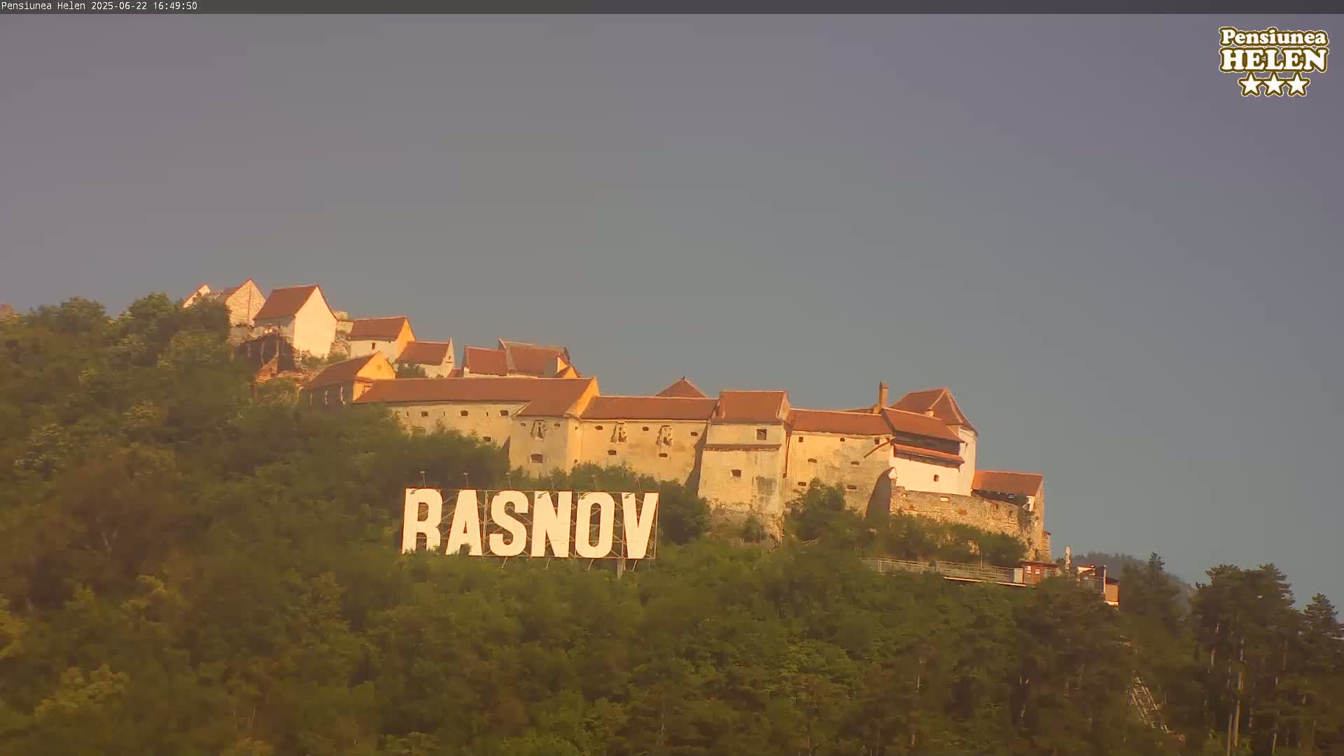 A large stone castle sits atop a tree-covered hill, with a sign reading "RASNOV" in the foreground under a clear, light-grey sky.