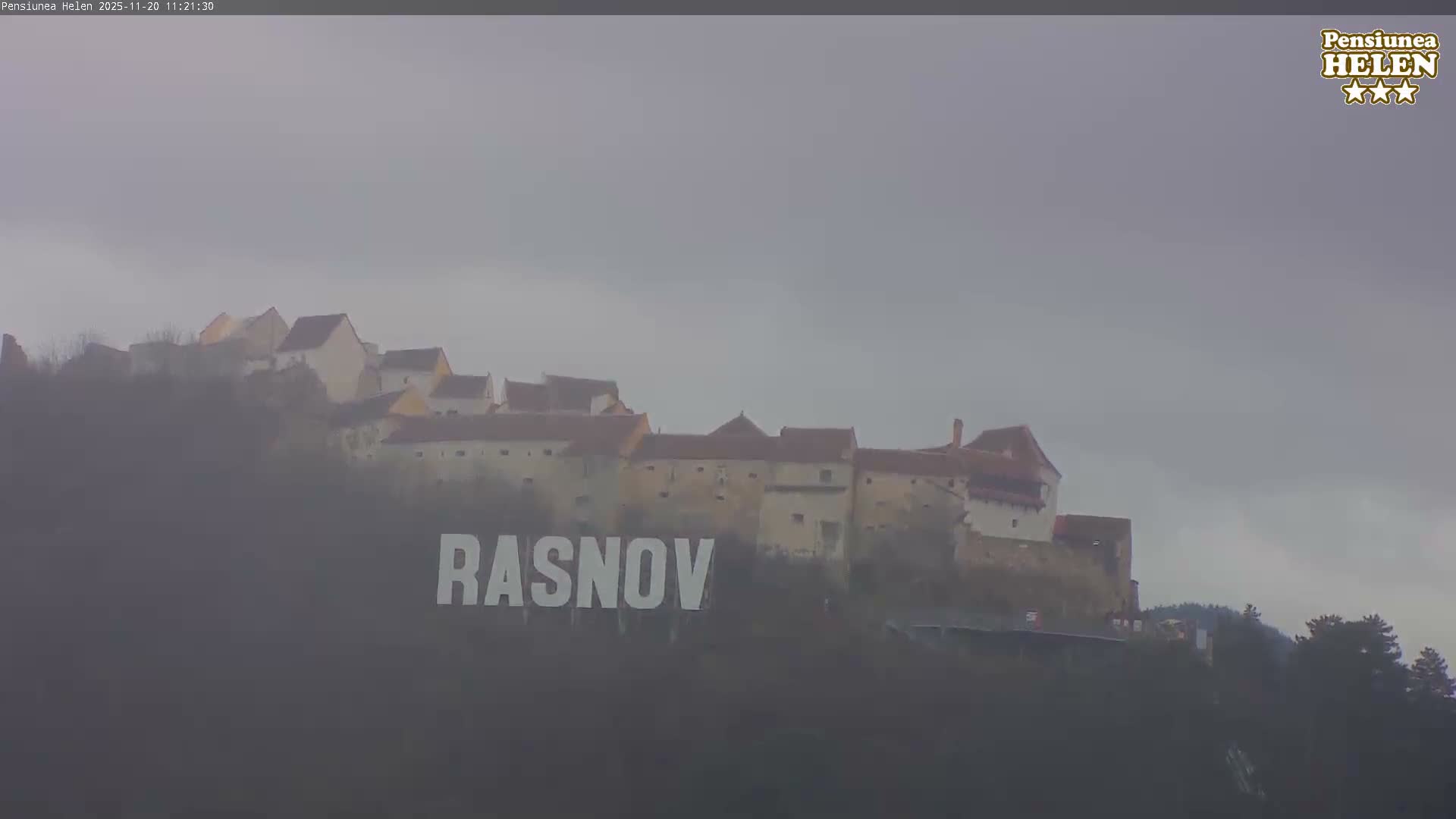 A historic fortified town with reddish-roofed buildings is perched on a misty hill under an overcast sky, with fog obscuring parts of the landscape.