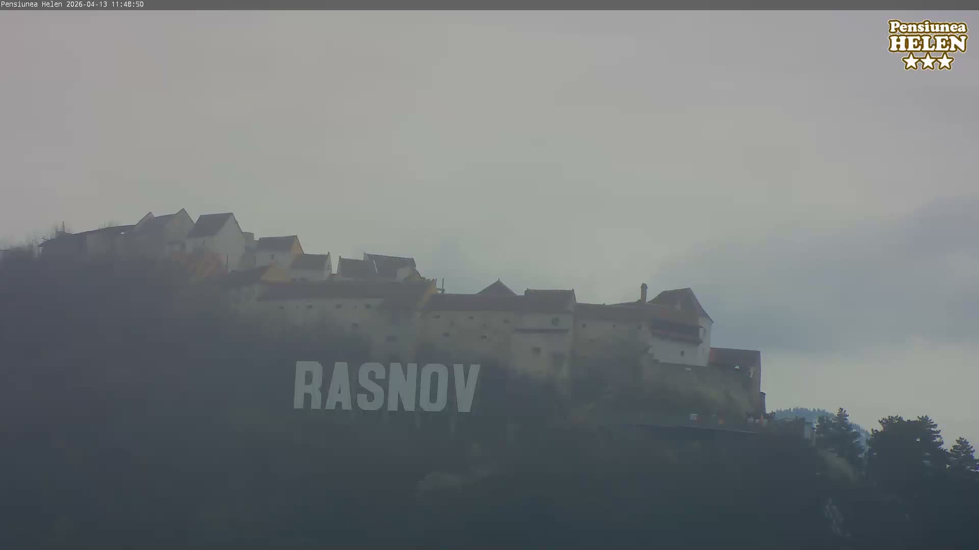 An ancient-looking fortified town or castle complex is perched on a misty hillside under a partly cloudy sky.