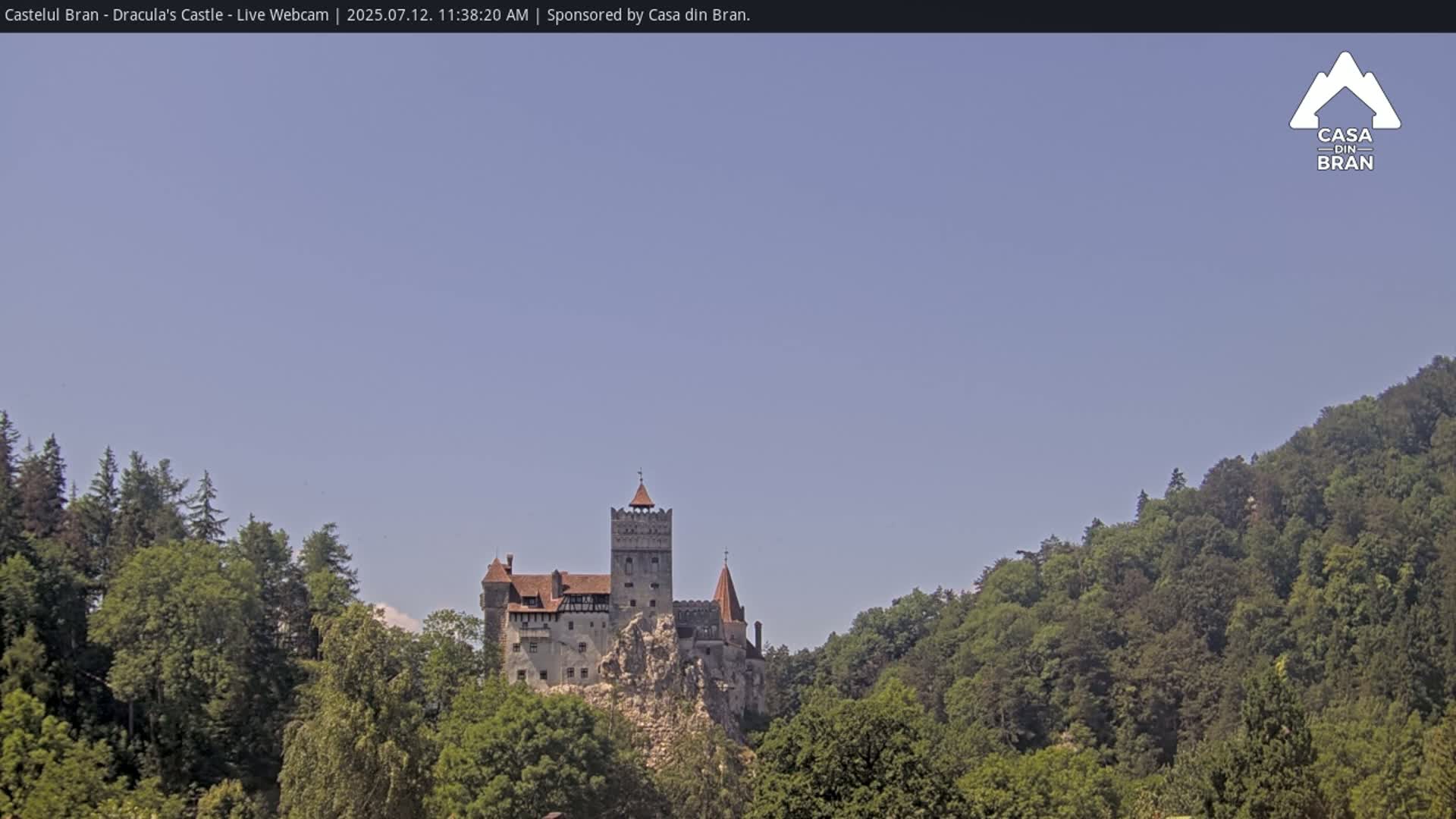 Bran Castle sits atop a rocky outcrop surrounded by lush green forests under a clear blue sky.