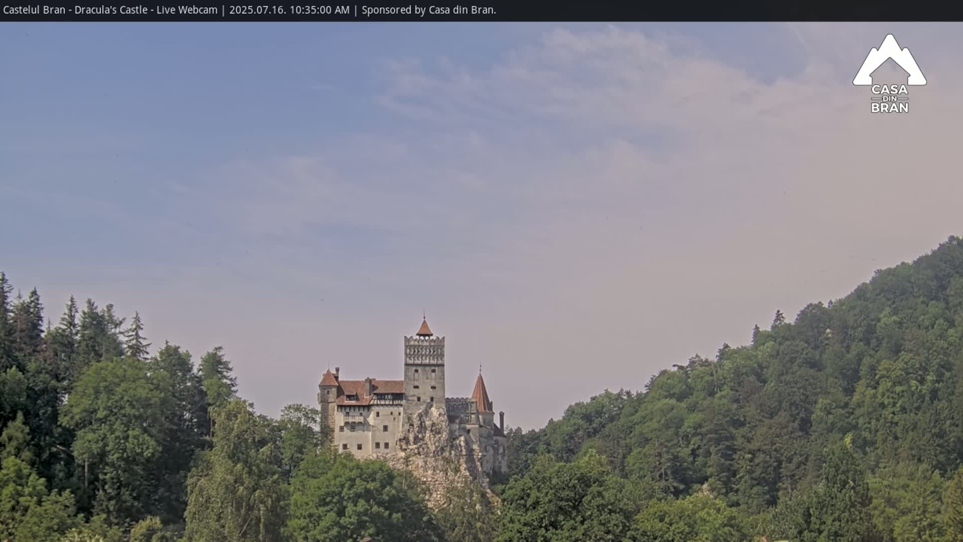 A medieval castle sits atop a rocky outcrop, surrounded by lush green forests under a mostly clear, light blue sky.