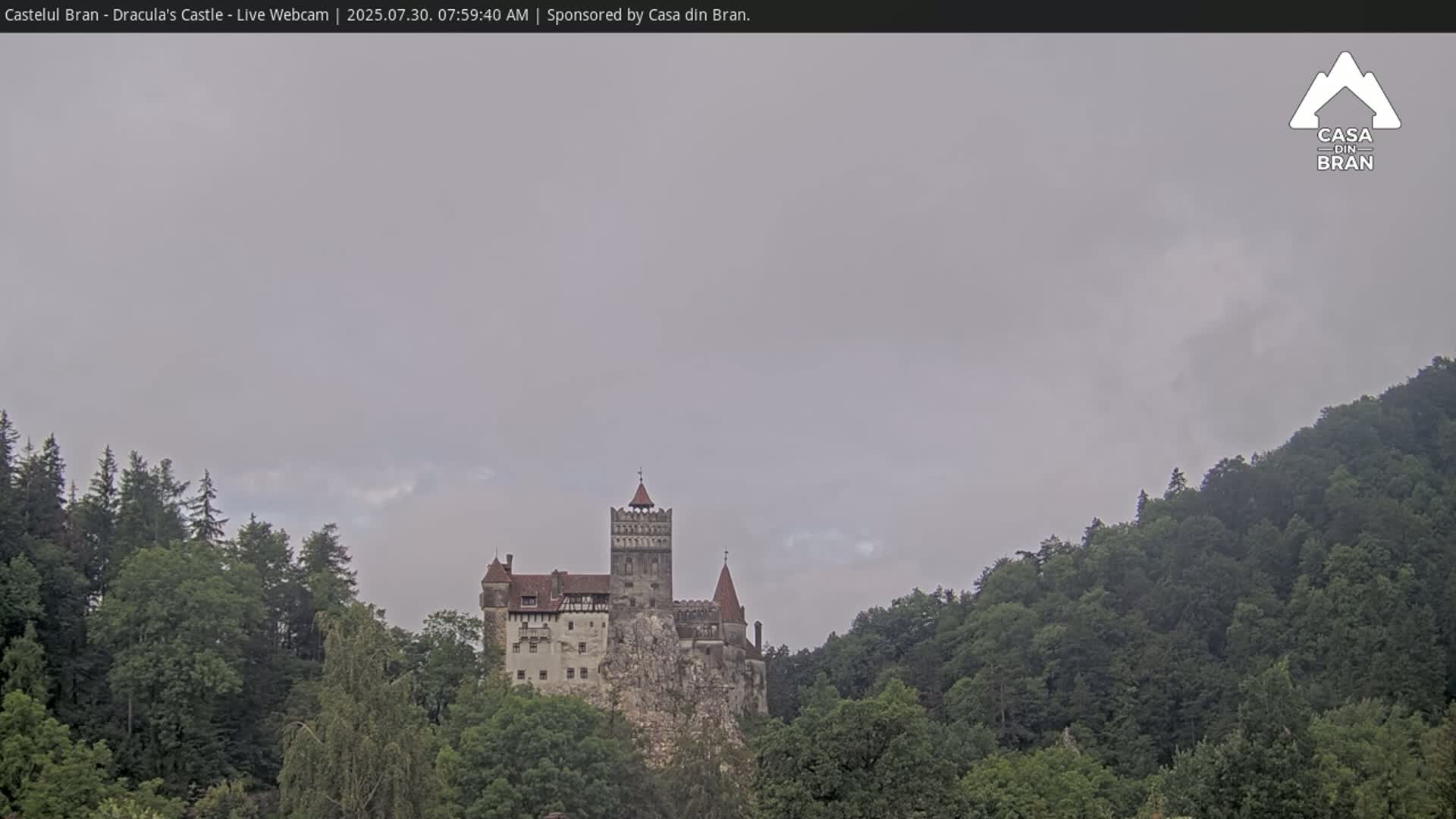 A medieval castle sits atop a hill covered in lush green trees under a partly cloudy sky.