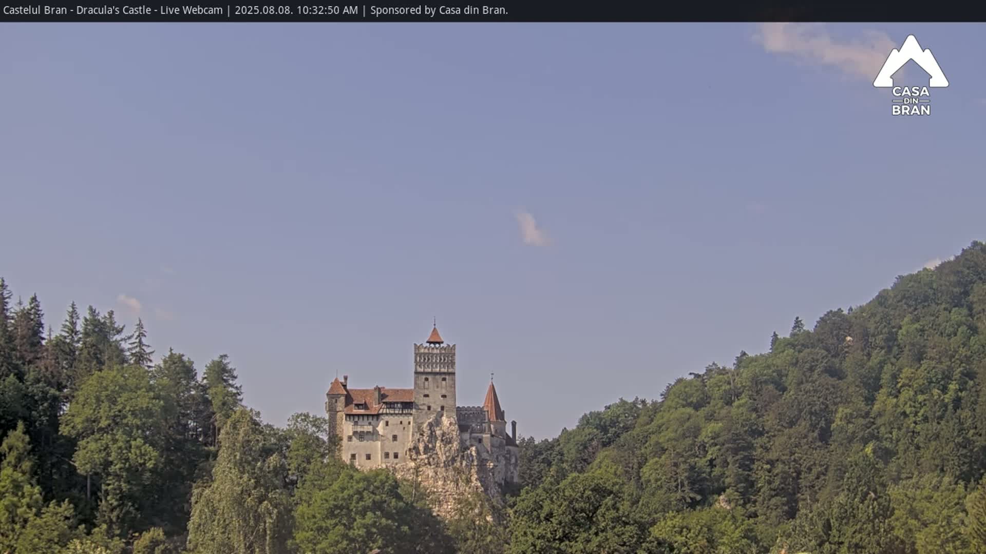 A large stone castle sits atop a cliff, surrounded by lush green trees under a mostly clear, light blue sky.