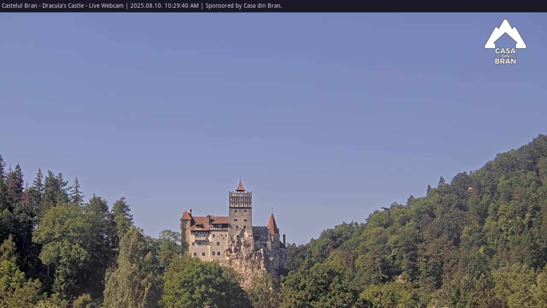 A stone castle sits atop a rocky cliff, surrounded by lush green forests under a clear blue sky.