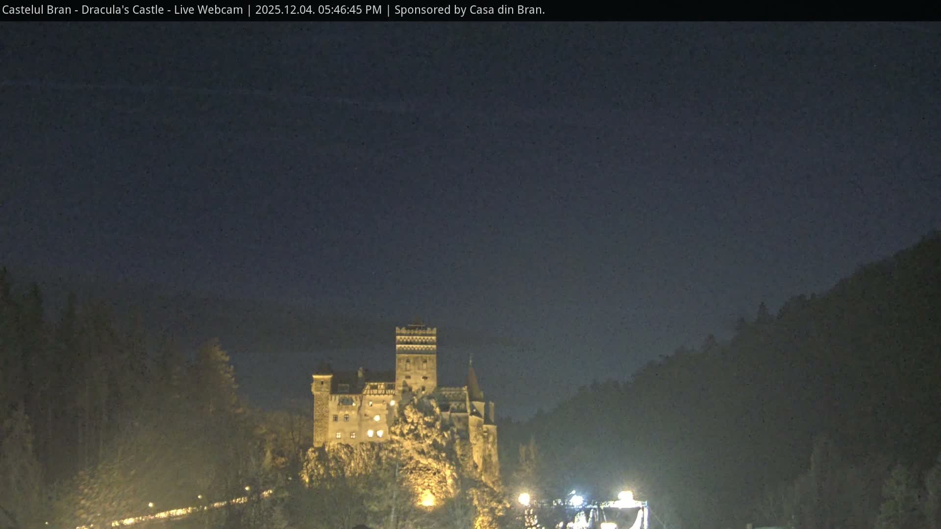 An illuminated castle sits dramatically atop a rocky hill, surrounded by dark, tree-covered slopes under a hazy night sky.