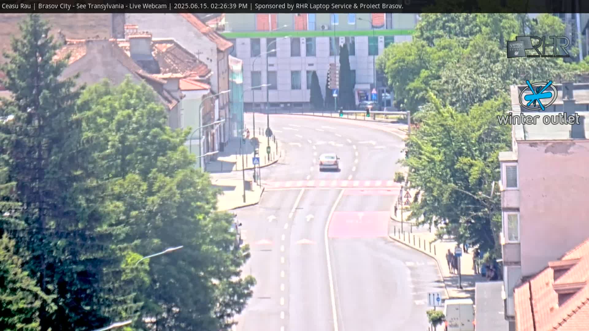 A sunny day shows a curved road with light traffic passing through a town with buildings and lush green trees lining the streets.