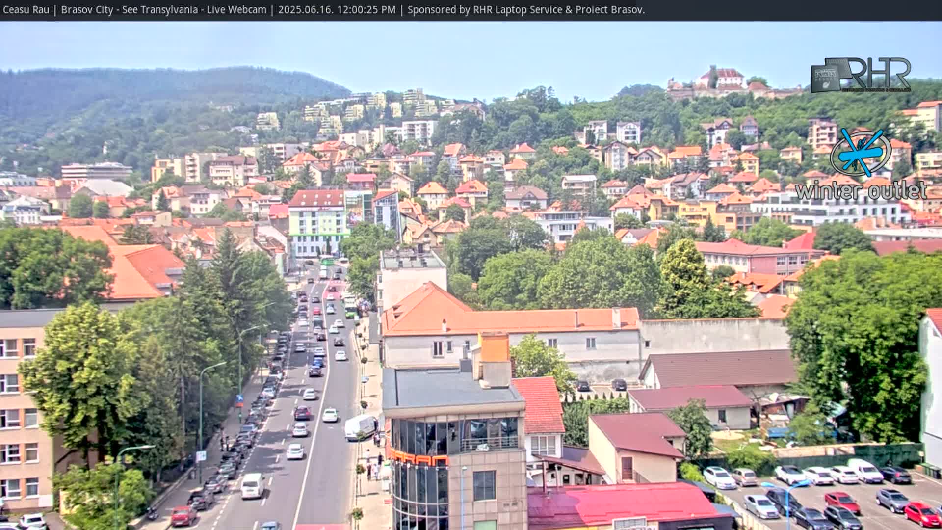 A sunny day overlooking a city with many buildings, lush green trees, and a busy road with cars.