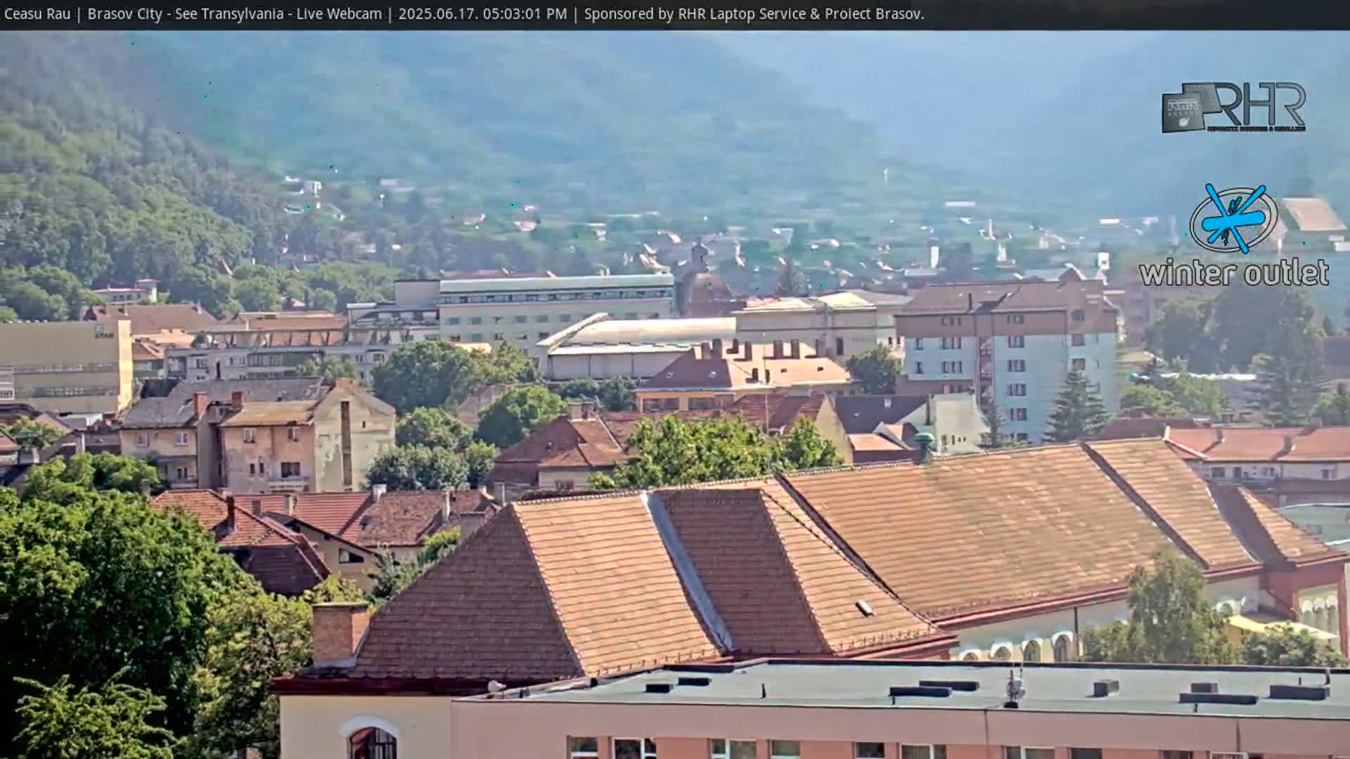 A hazy, sunny view overlooks a city of European-style buildings with red-tiled roofs, nestled amongst green trees and hills.