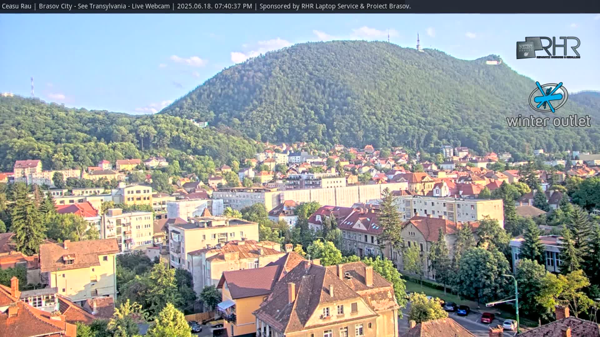 A high-angle, long shot of a city nestled beneath a large, green hill on a partly sunny day.