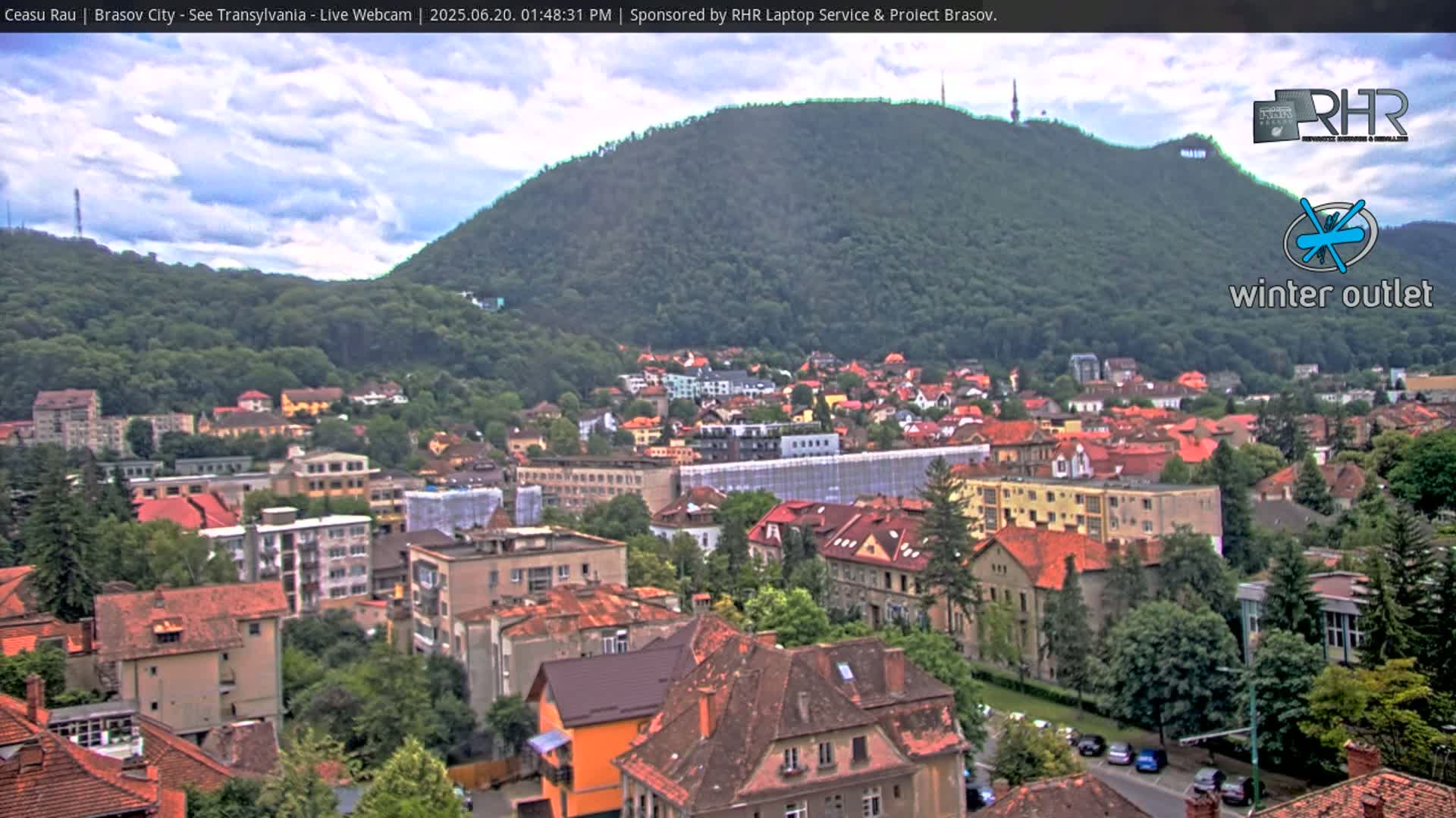 A partly cloudy day overlooks a city nestled at the base of a large, forested hill.