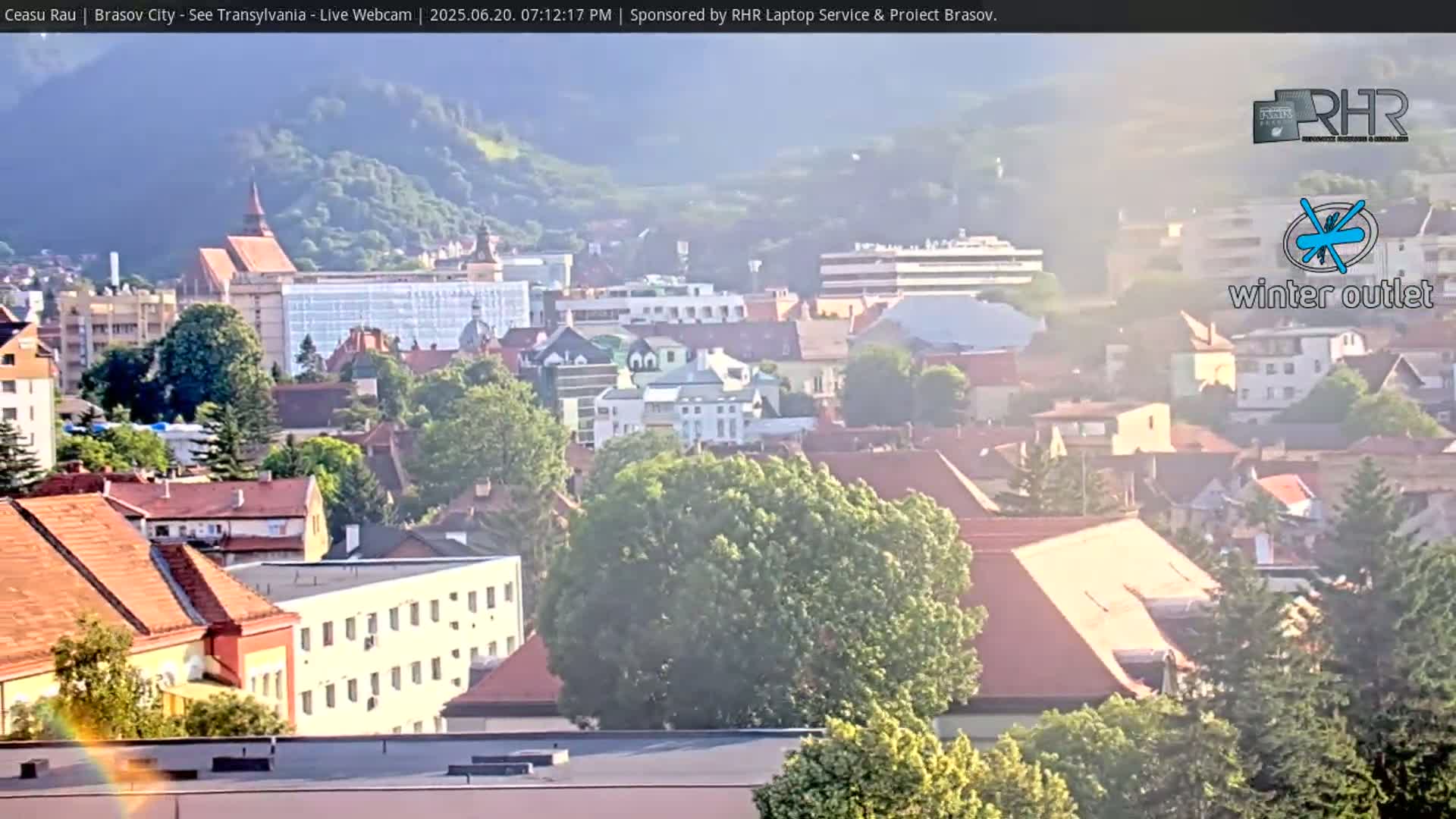 A hazy, sunny view of a city with numerous red-tiled roofs and buildings nestled amongst green trees and hills.