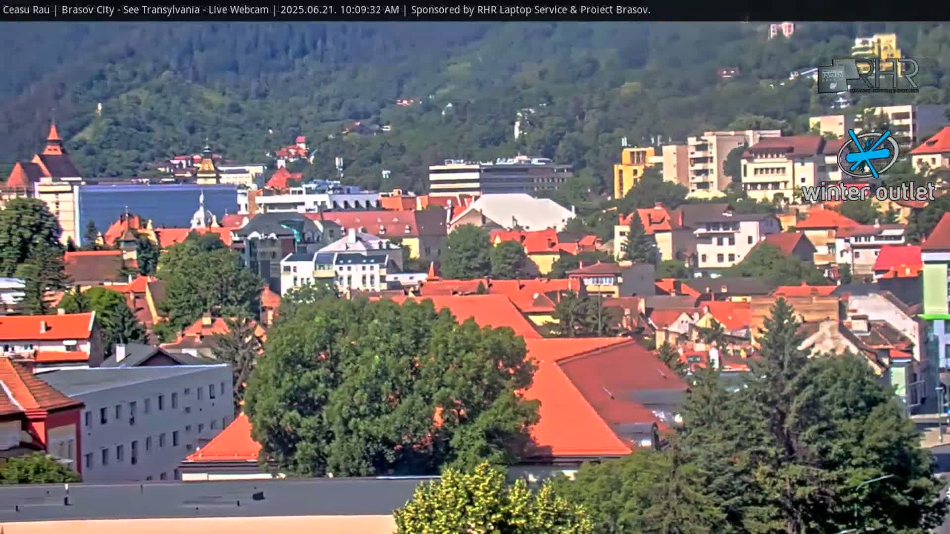 A sprawling cityscape of red-roofed buildings nestled amongst green hills on a partly sunny day.