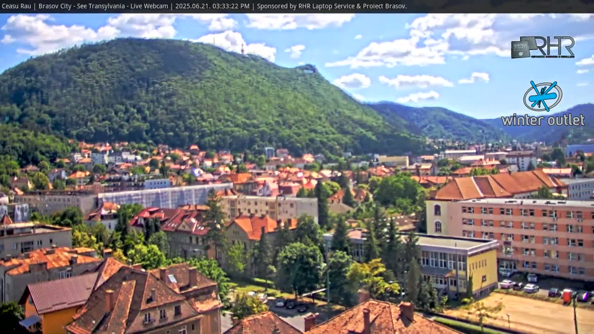 A sprawling town nestled beneath a lush green mountain, under a partly sunny sky.