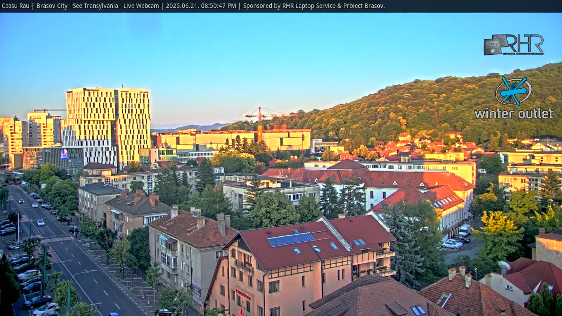 A city street scene at sunset shows a mix of modern and older buildings, with a hill covered in green trees in the background.