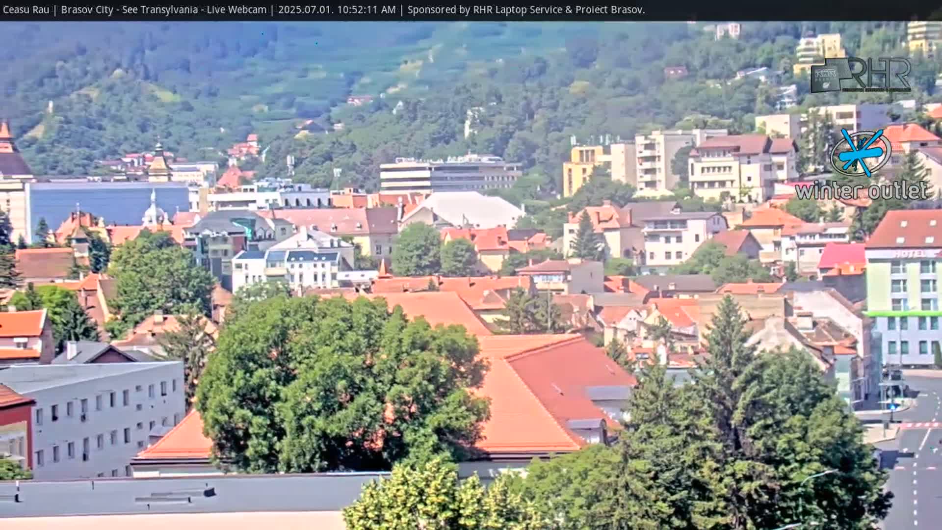 A sunny day reveals a cityscape of numerous red-roofed buildings nestled amongst green trees, situated on a hillside.