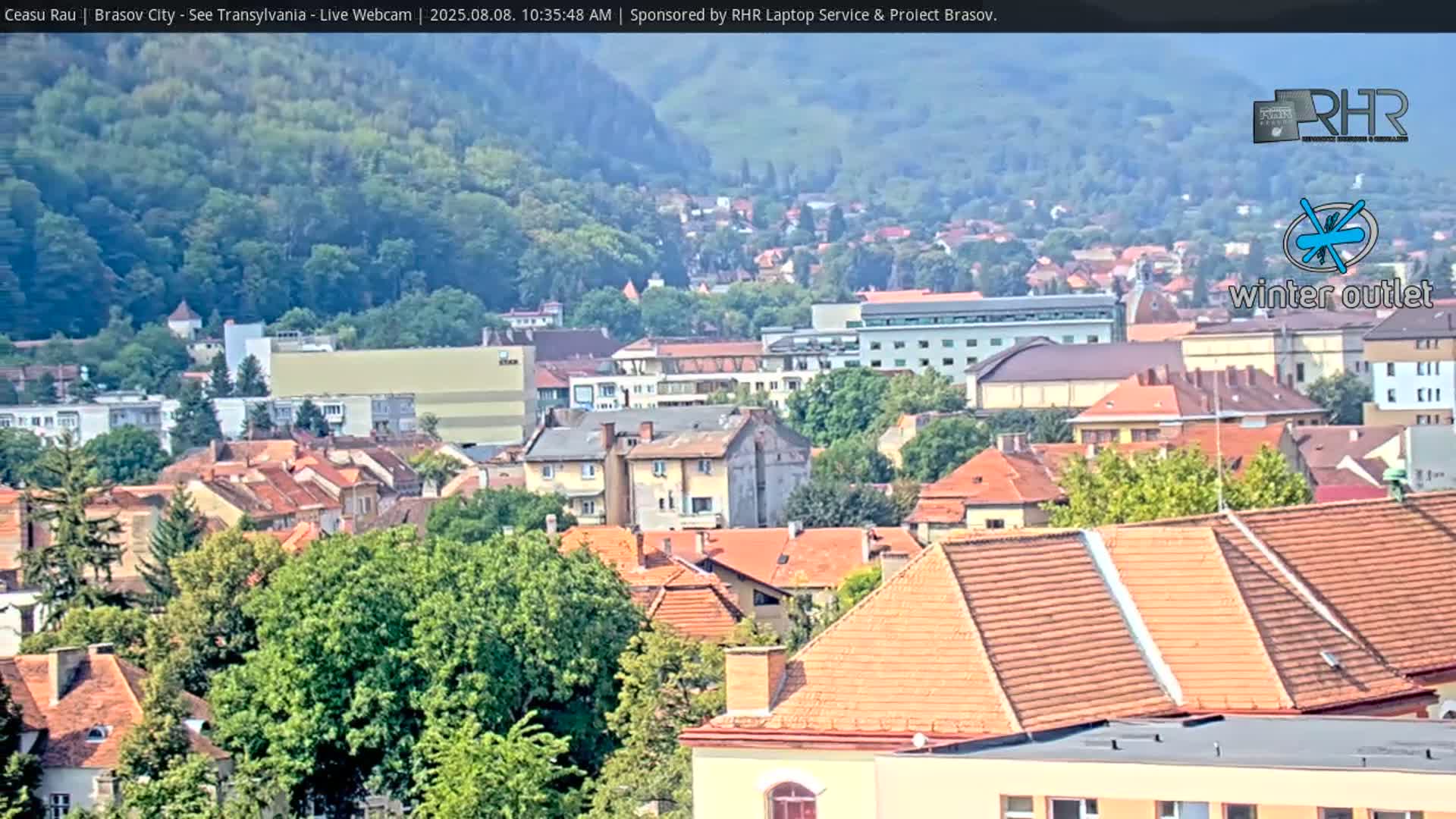 A long shot of a city nestled in a valley shows many buildings with orange-tiled roofs, green trees, and a hazy sky.