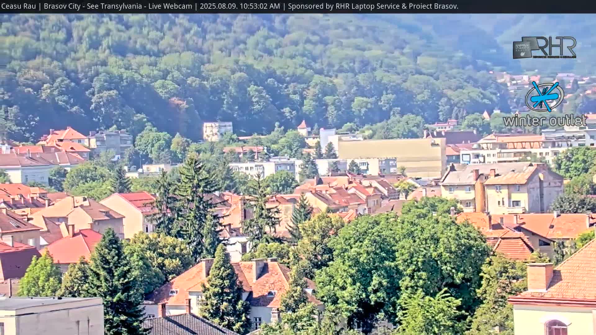A sunny view of a city with red-tiled rooftops, green trees, and a hill in the background.