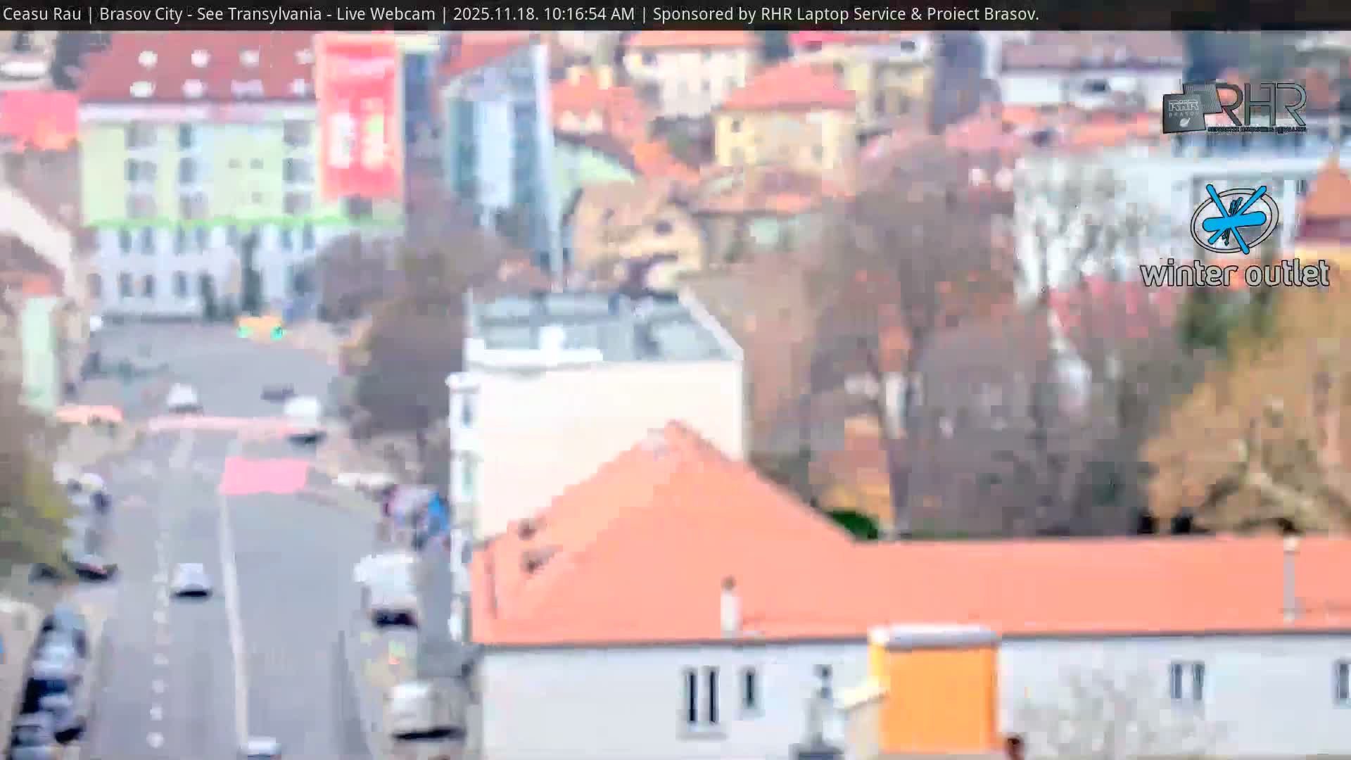 A sunny city street scene shows light traffic moving along a multi-lane road, with pedestrians walking on a sidewalk lined with buildings and trees.