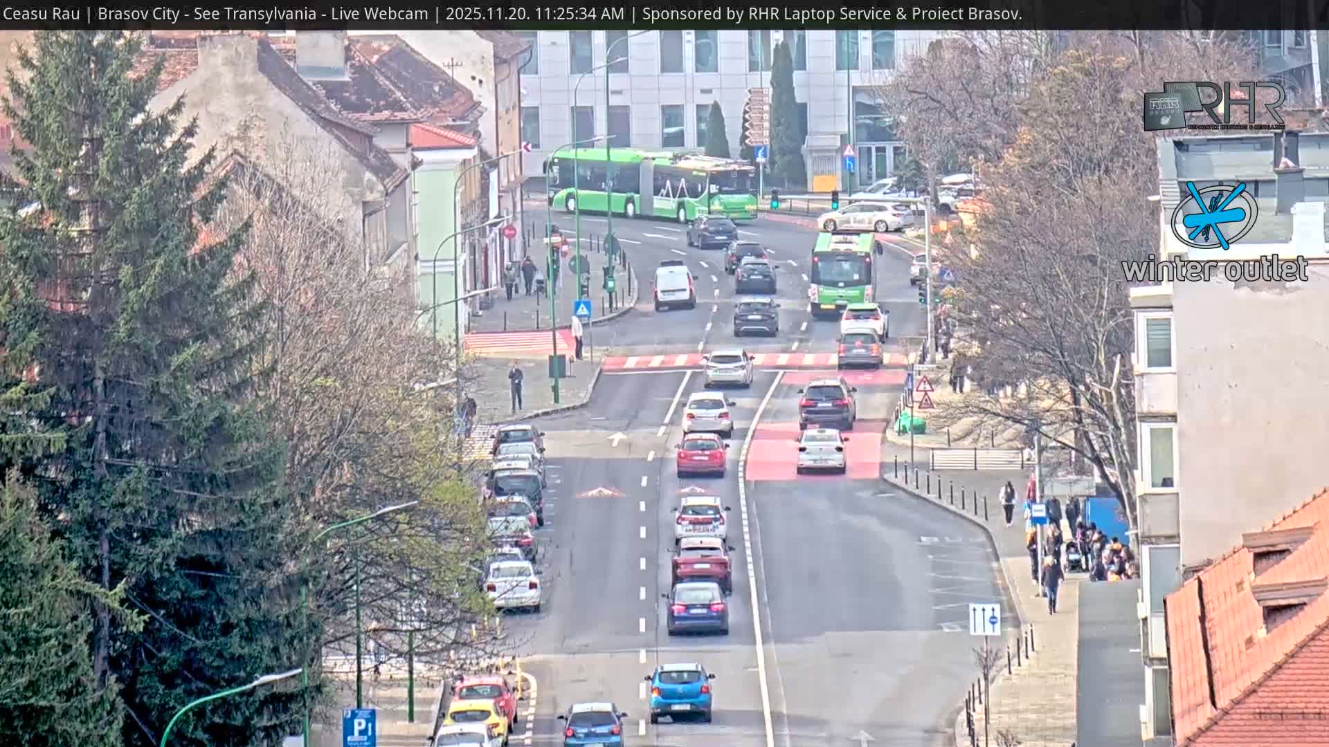 A bustling city street, featuring multiple lanes of traffic with cars and green buses, is flanked by sidewalks with pedestrians, bare deciduous trees, and a mix of traditional and modern buildings under a clear sky.