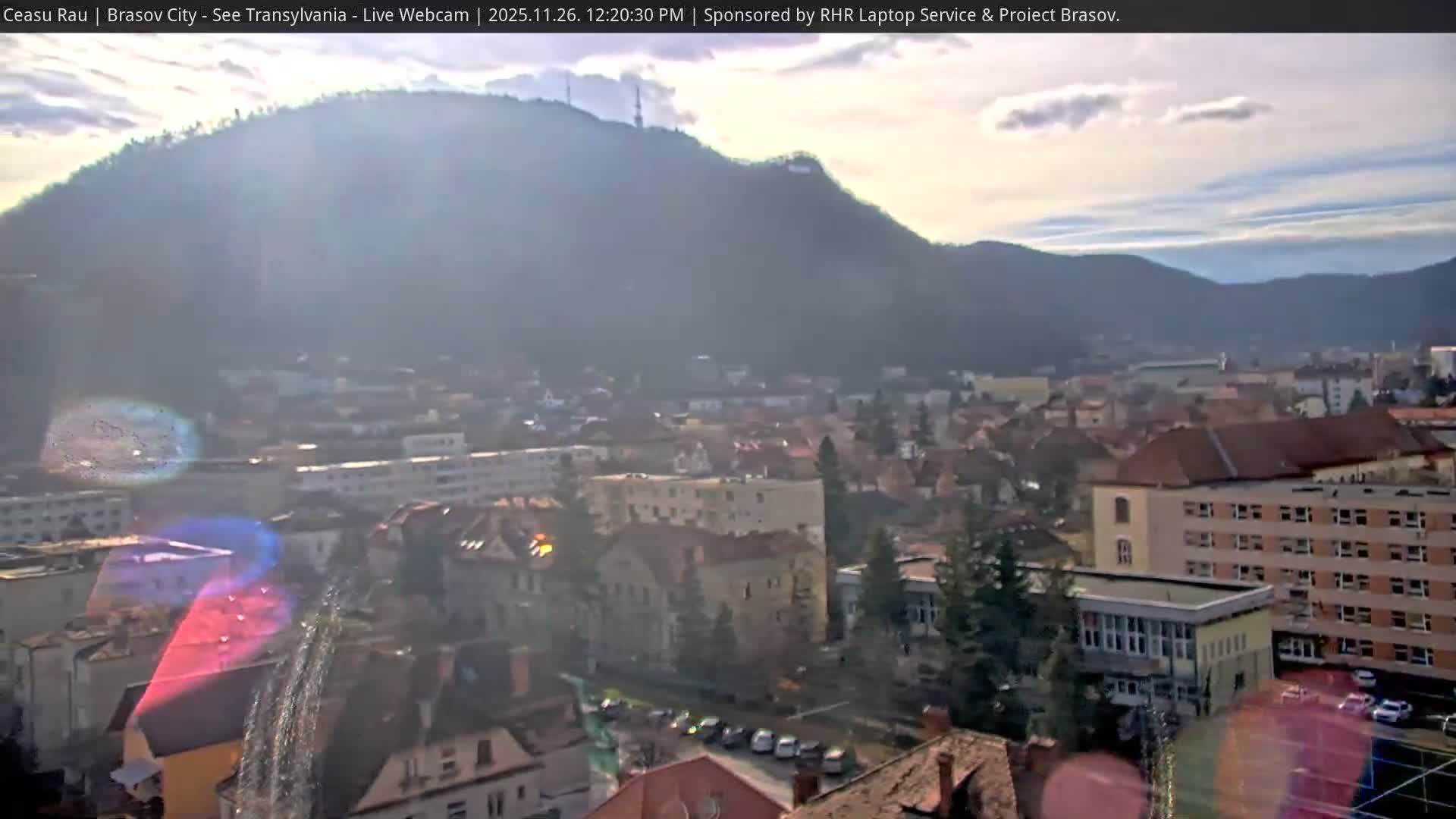 A city with a mix of buildings and scattered trees is nestled against large, forested mountains under a bright, partly cloudy sky with significant sun glare and atmospheric haze.