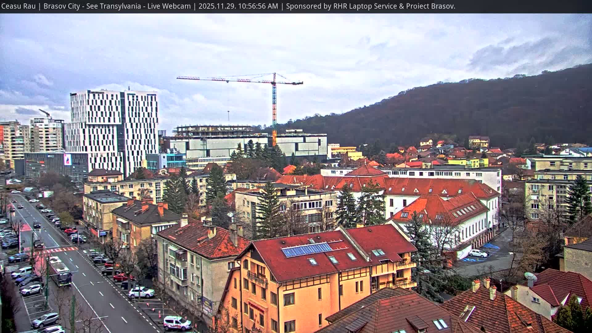 On an overcast day, an urban landscape featuring a mix of modern high-rises, traditional red-roofed houses, and active construction cranes sprawls before a tree-covered mountain.