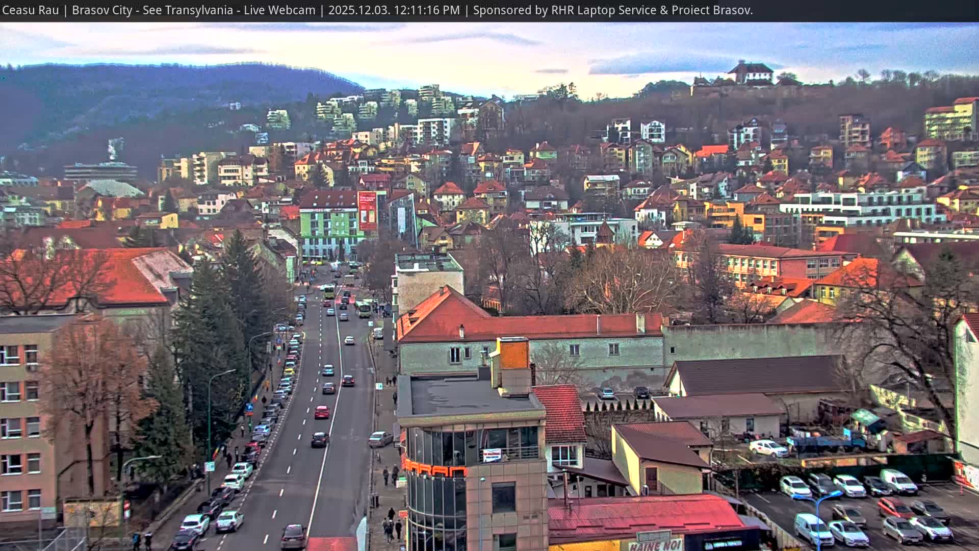 An elevated view captures a busy urban street with traffic and many buildings, stretching towards a city nestled against distant hills under an overcast daytime sky, with mostly bare trees suggesting a cool season.