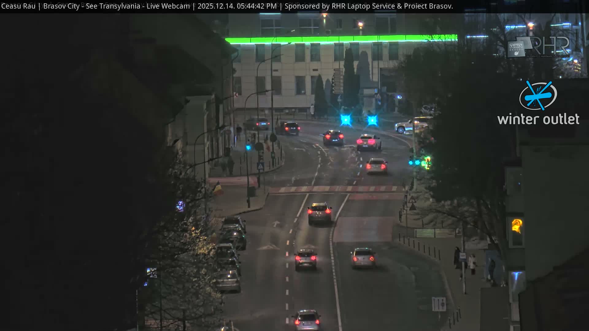 An elevated view captures a busy urban street with traffic and many buildings, stretching towards a city nestled against distant hills under an overcast daytime sky, with mostly bare trees suggesting a cool season.