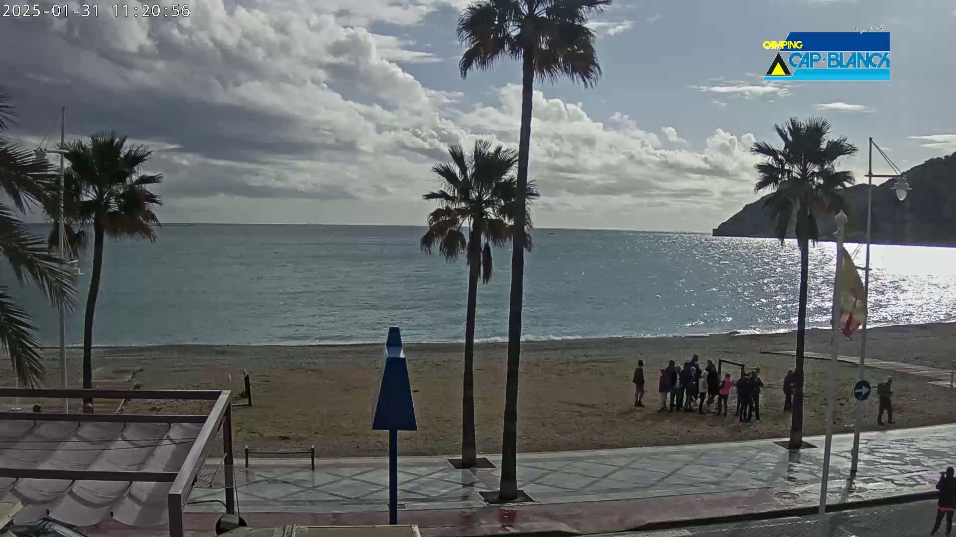 A mostly cloudy day shows a beach scene with palm trees, people gathered on the sand, and a calm sea in the background.
