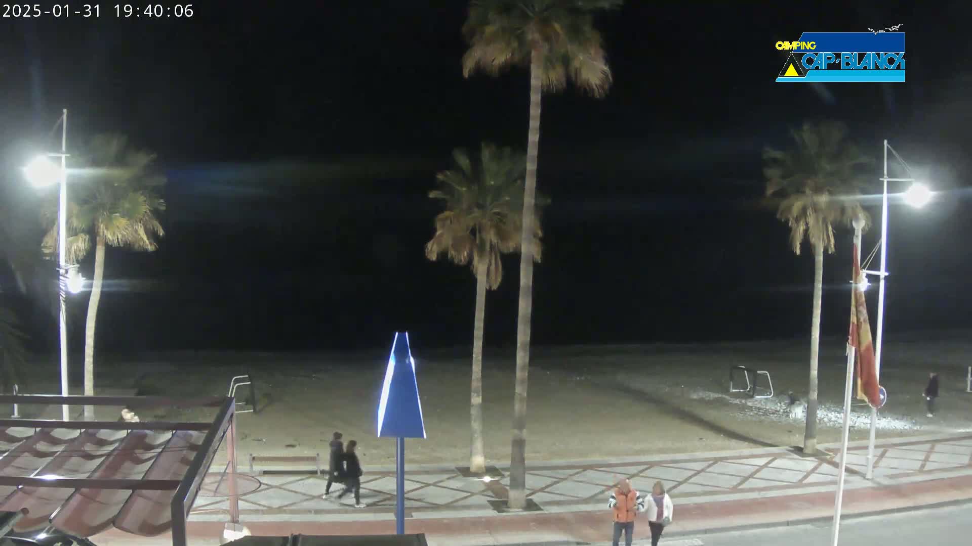 At night, a sandy beach is visible with palm trees lining a paved walkway where several people are walking.