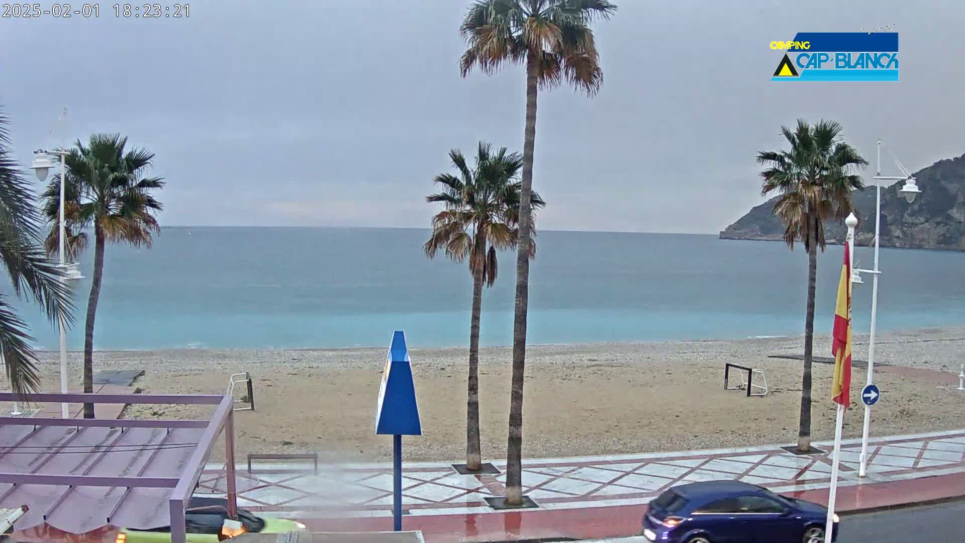 A rain-swept beach scene shows palm trees lining a promenade next to a sandy beach and calm turquoise water under an overcast sky.