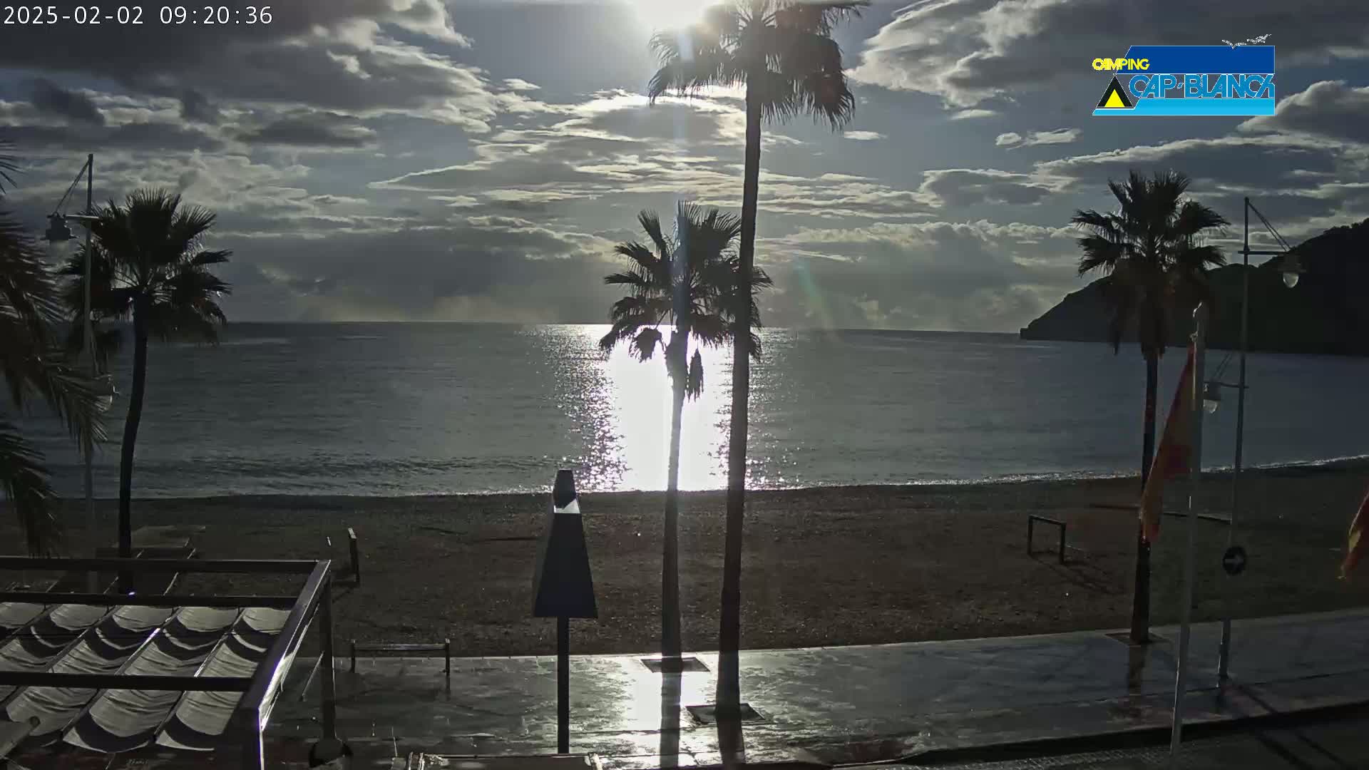A partly cloudy sunrise illuminates a beach with palm trees, a calm sea, and a paved walkway.