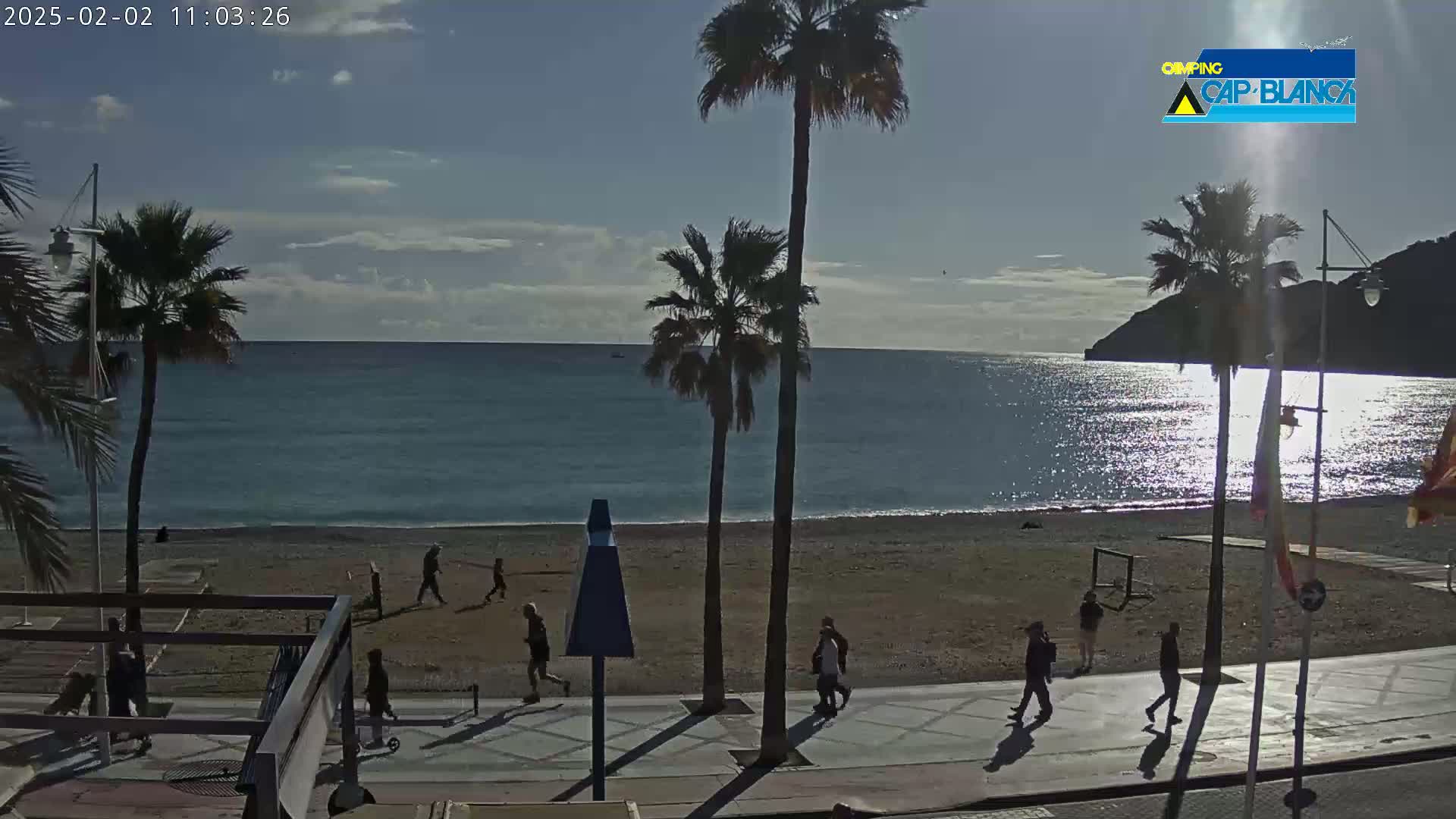 A mostly sunny day at a beach, showing palm trees, people walking on a paved walkway next to a sandy beach and calm ocean water.