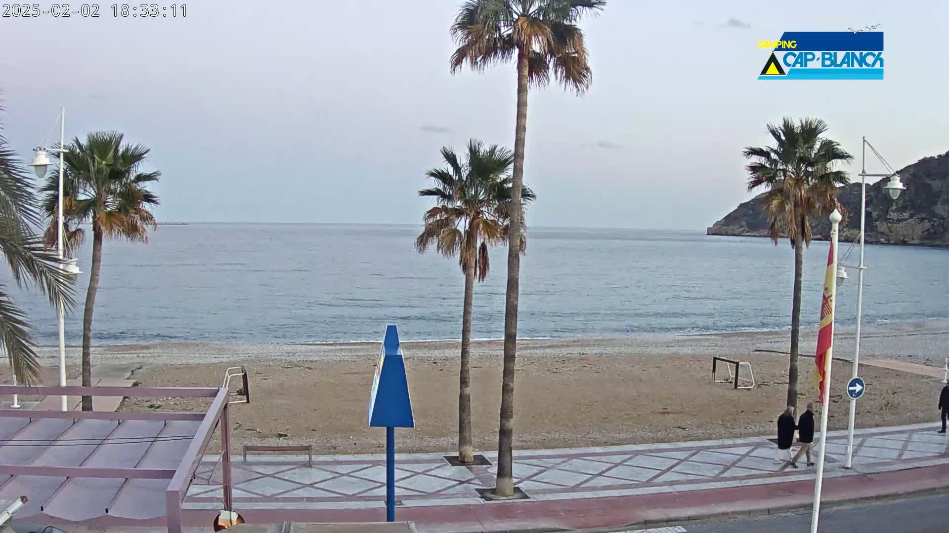 A calm, overcast day reveals a sandy beach with palm trees lining a promenade where two people stroll, overlooking a tranquil sea and distant hills.