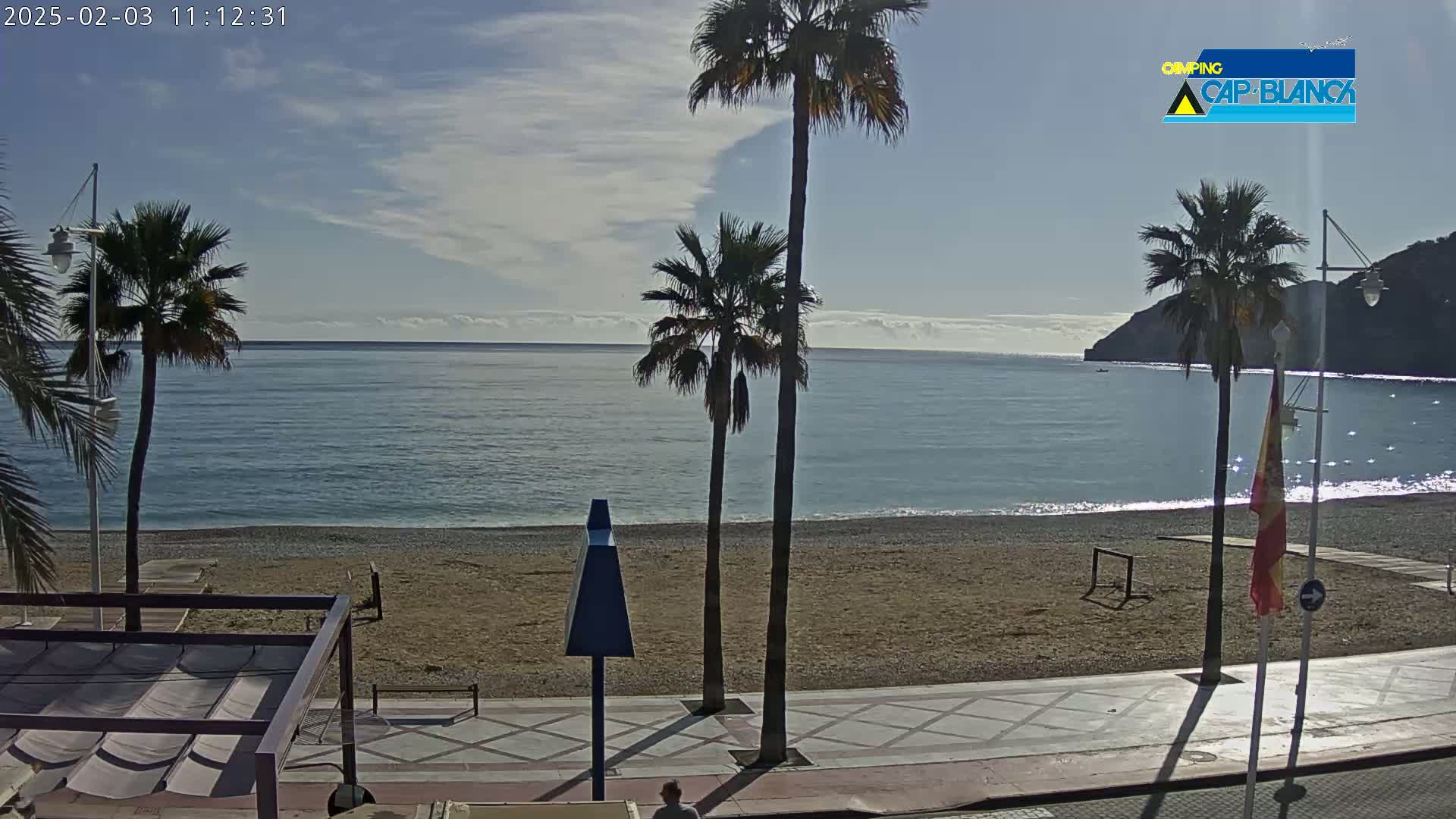 A mostly sunny beach scene with calm blue water, palm trees, and a paved walkway.